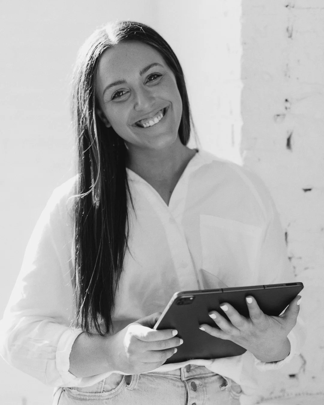 A smiling woman holding a tablet, standing near a brick wall, black and white photo.
