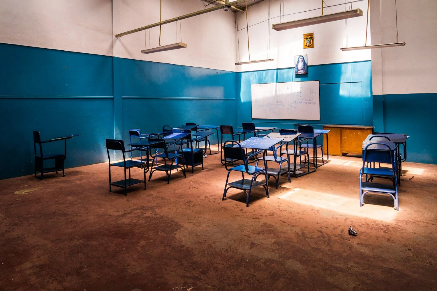 An empty classroom with blue walls, a whiteboard, a small wooden desk, and scattered chairs and desks. There is a portrait and a religious picture hanging above the whiteboard. The floor appears worn and there is a single small object on the floor.