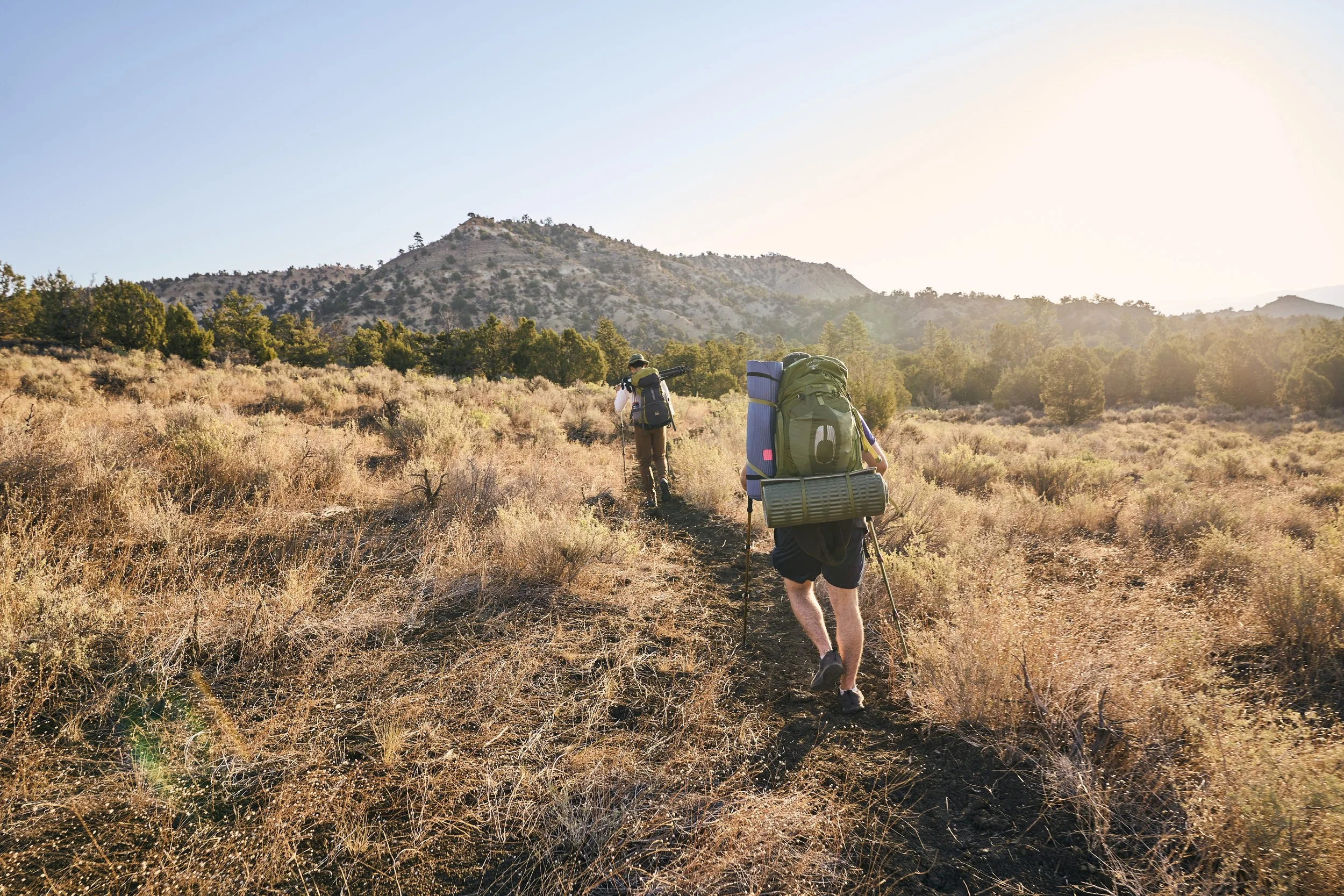 Two hikers with large backpacks walking on a dirt trail through dry grass in a mountainous area at sunset.