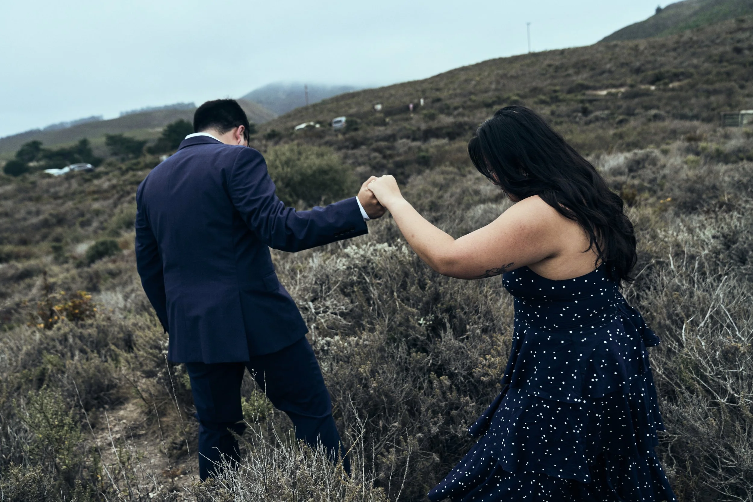 A man in a suit and a woman in a polka dot dress holding hands in a hillside landscape.