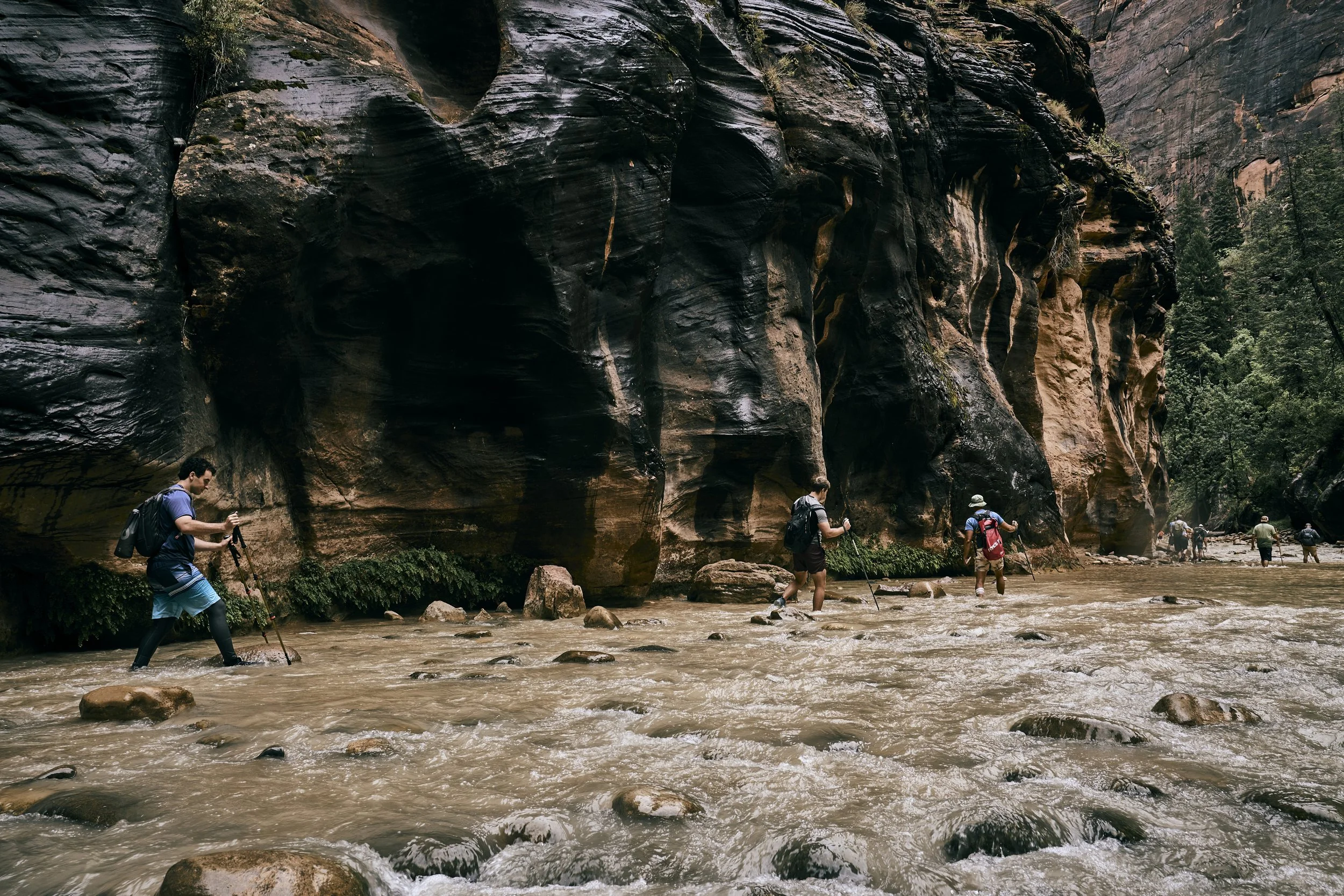 People hiking across a shallow river with large rocks, surrounded by tall rock formations and green trees in a canyon.