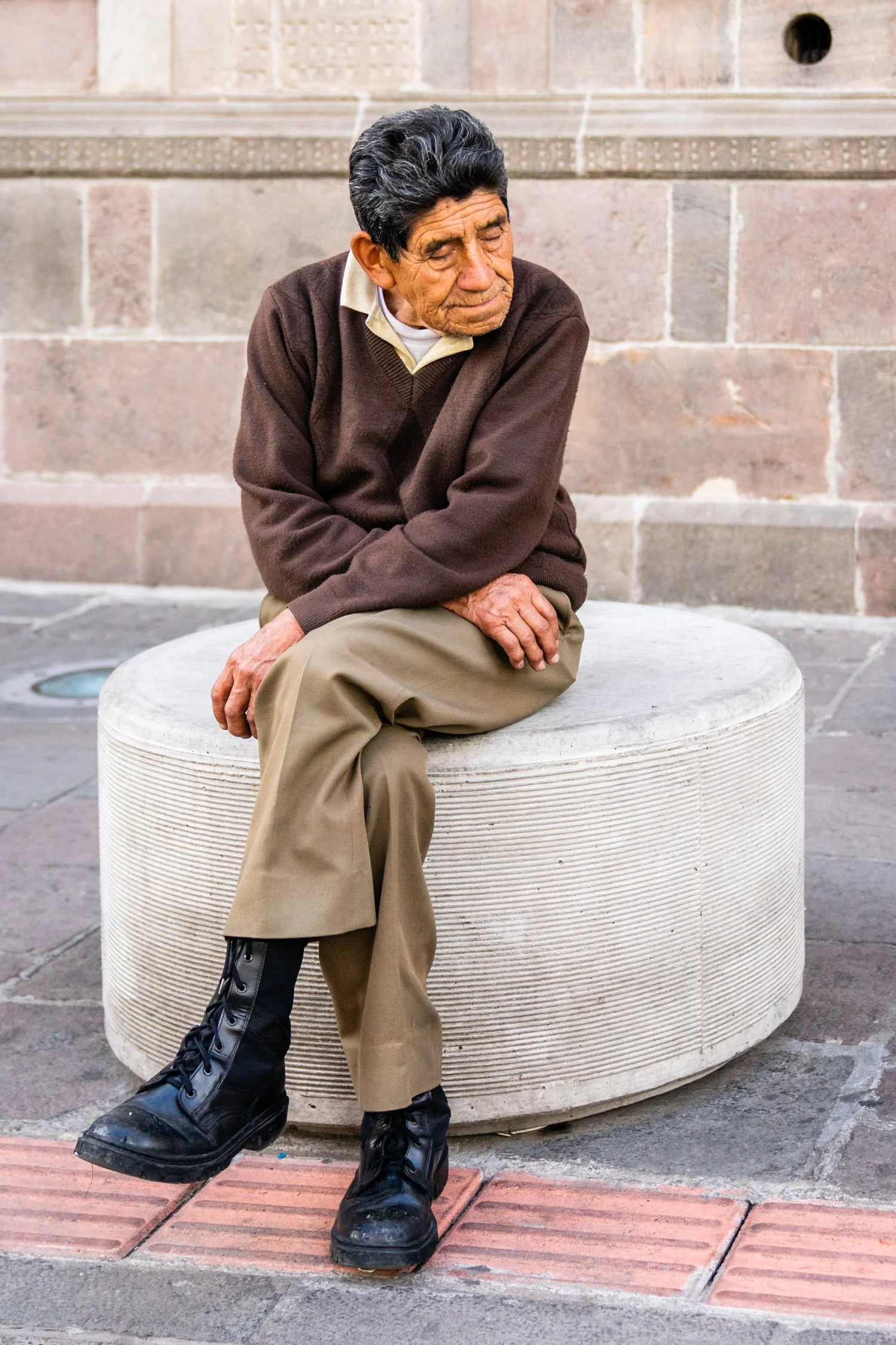 An elderly man with dark hair, wearing a brown sweater, beige pants, and black boots, sitting on a rounded stone bench outdoors against a stone wall, with one leg crossed over the other.