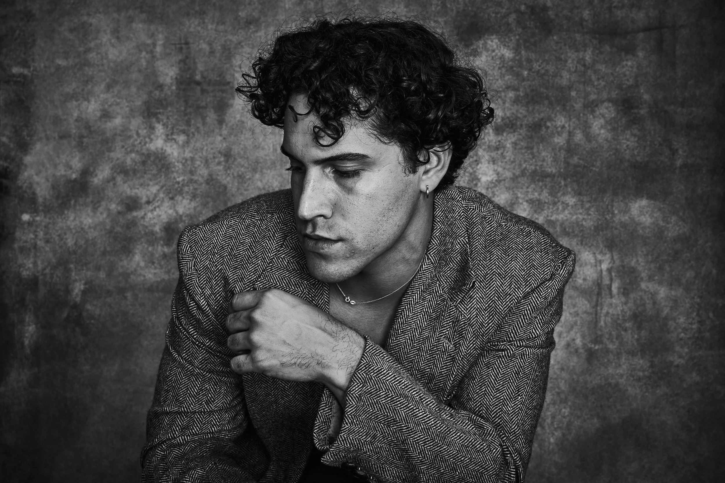 Black and white portrait of a young person with curly hair, wearing a patterned blazer, looking down with a contemplative expression, against a textured background.