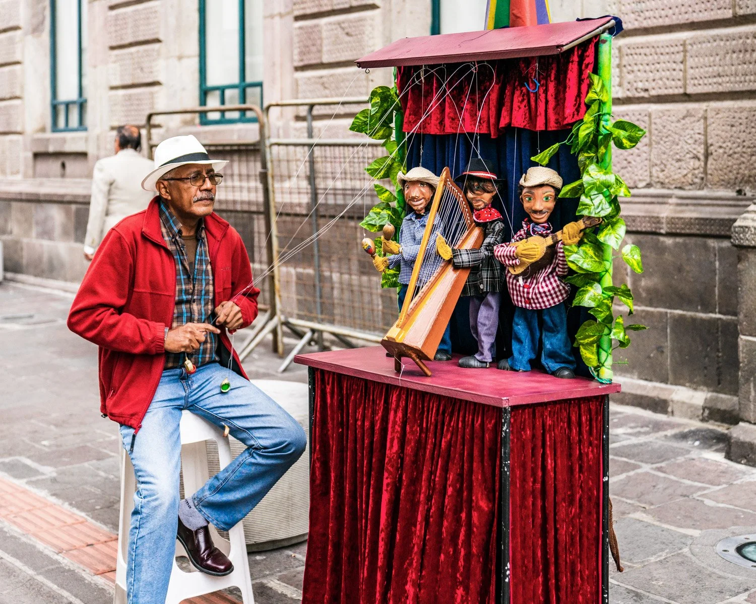 A man wearing a white fedora, red jacket, and glasses is operating a puppet show with marionette puppets on a small stage on a city street.