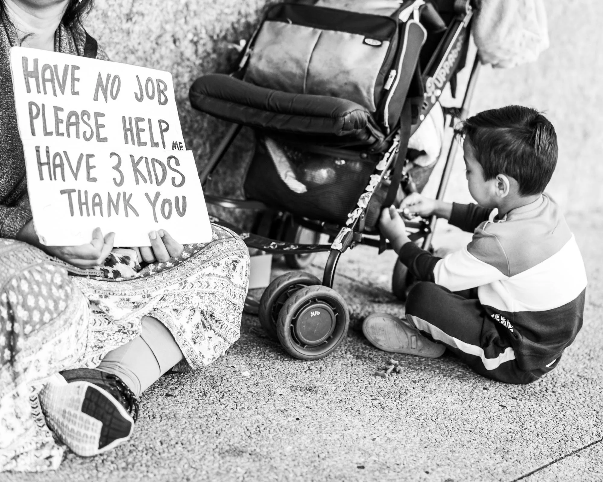 A woman holding a sign that says, "Have no job, please help me, have 3 kids, thank you." Sitting on the ground, next to a young boy who is crouching and looking at the ground, with a stroller and some bags nearby.