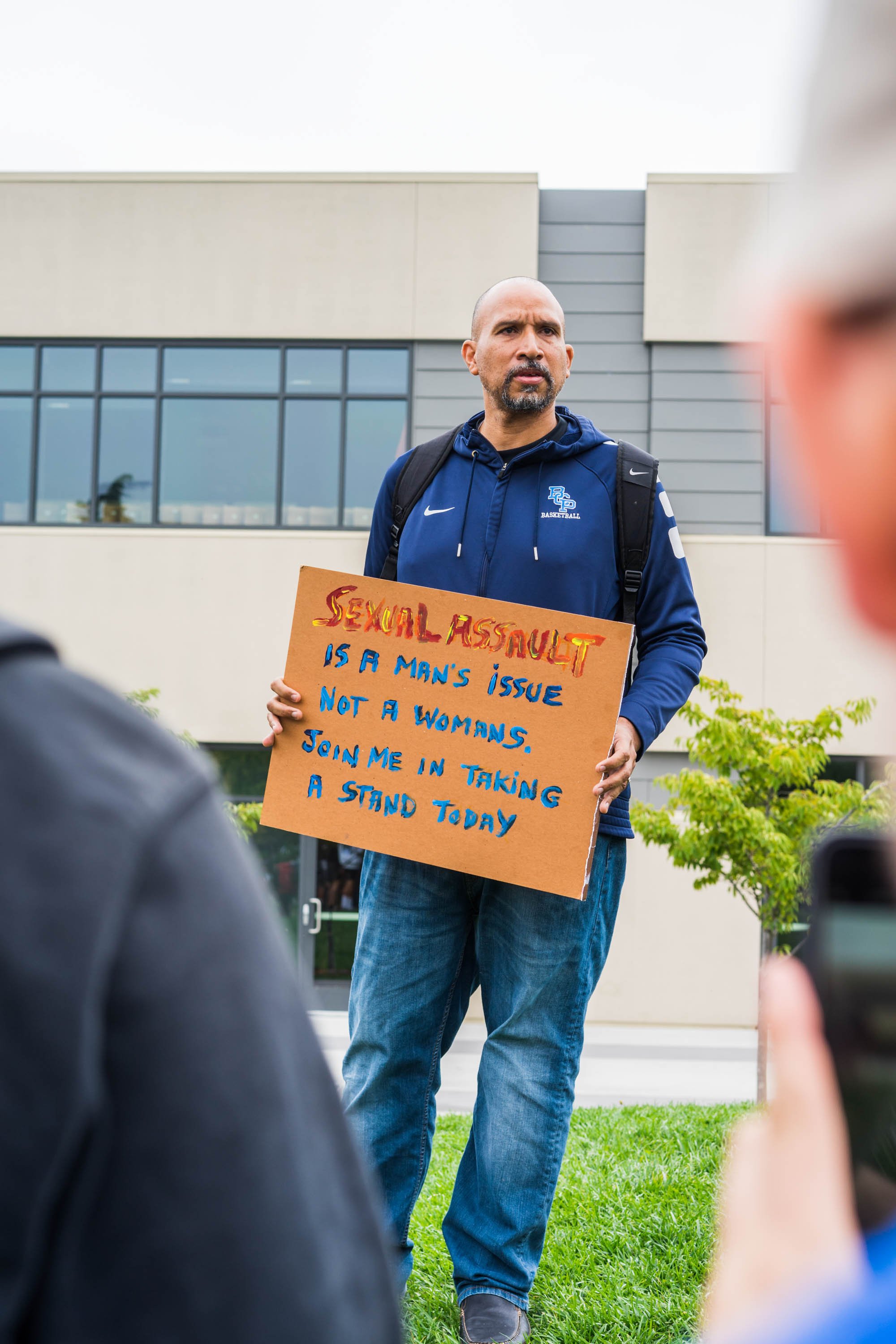 A man holding a protest sign that reads, 'Sexual assault is a man's issue, not a woman's. Join me in taking a stand today,' stands outdoors near a modern building with large windows, while several people appear to be listening or observing.
