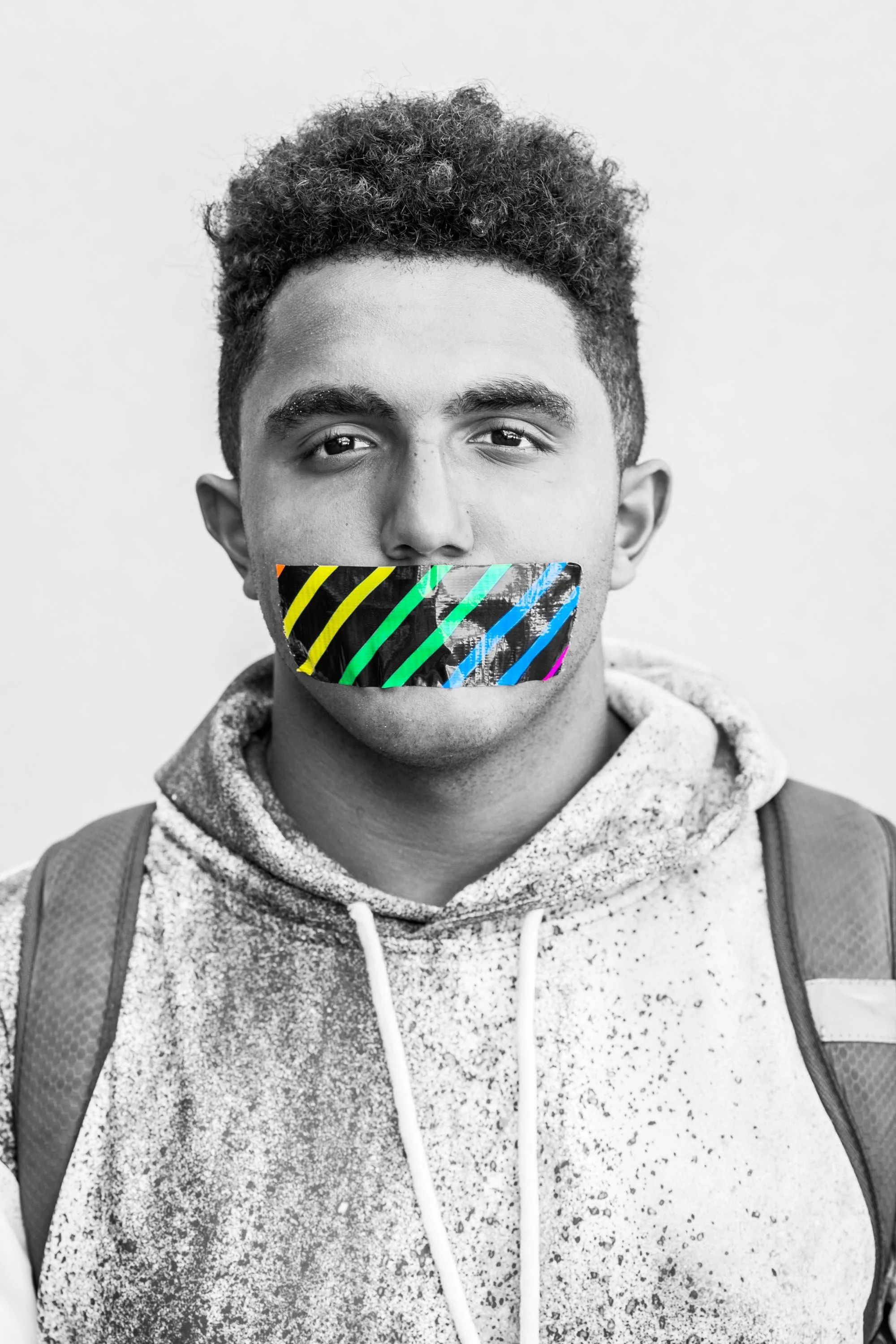 Young man with curly hair, wearing a hoodie and a backpack, with a colorful tape over his mouth in a black-and-white photo.