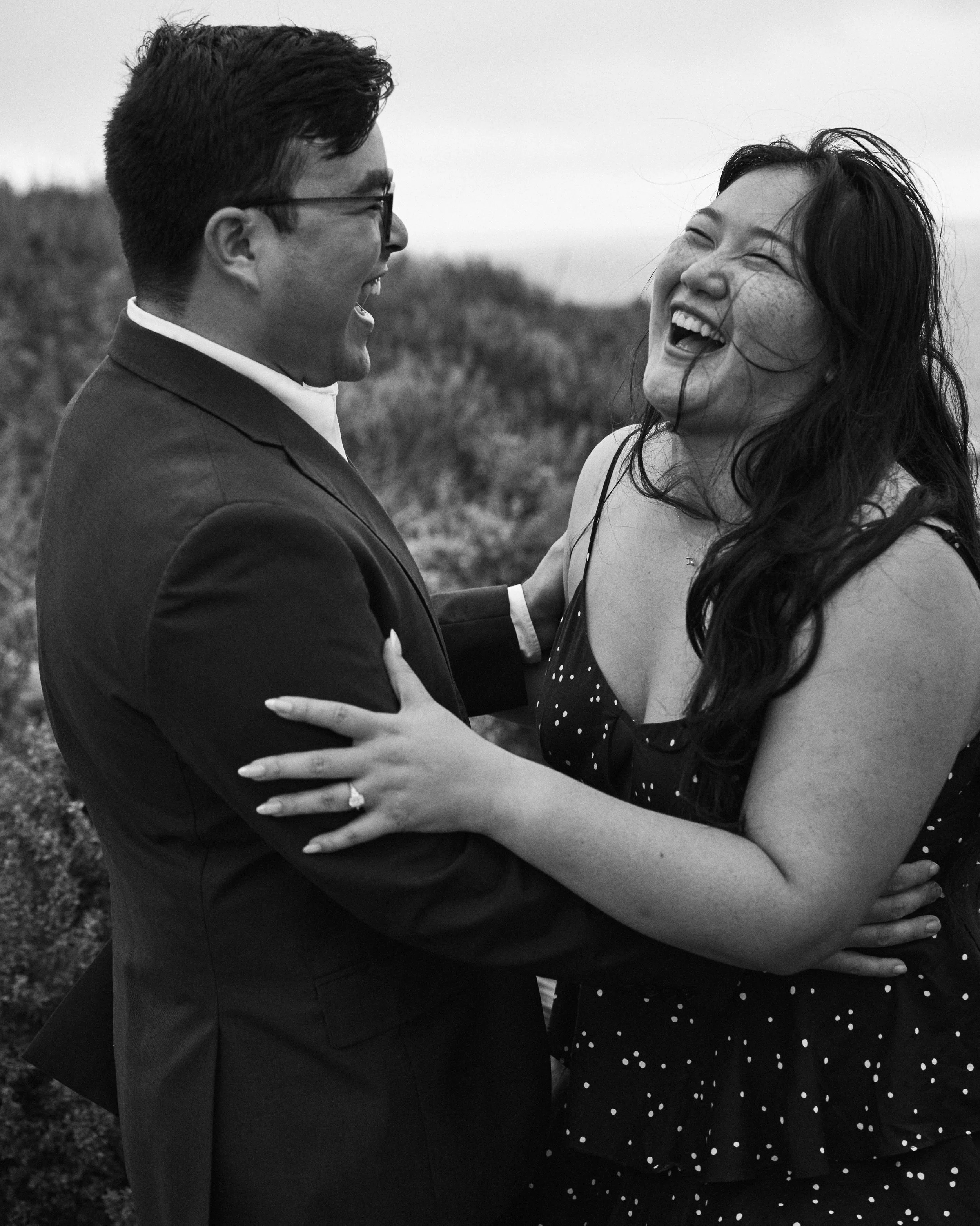 A black-and-white photo of a couple smiling and looking at each other. The man is wearing glasses and a suit, and the woman has long dark hair and is wearing a polka dot dress. They are holding each other in an outdoor setting with a blurred natural 