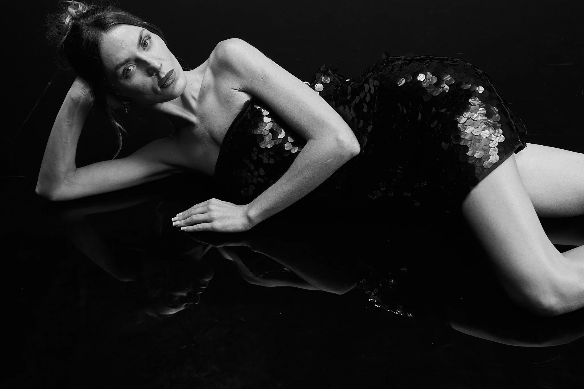 A woman with light skin and long hair, lying on a black reflective surface, wearing a sleeveless dress with sequins, looking directly at the camera.
