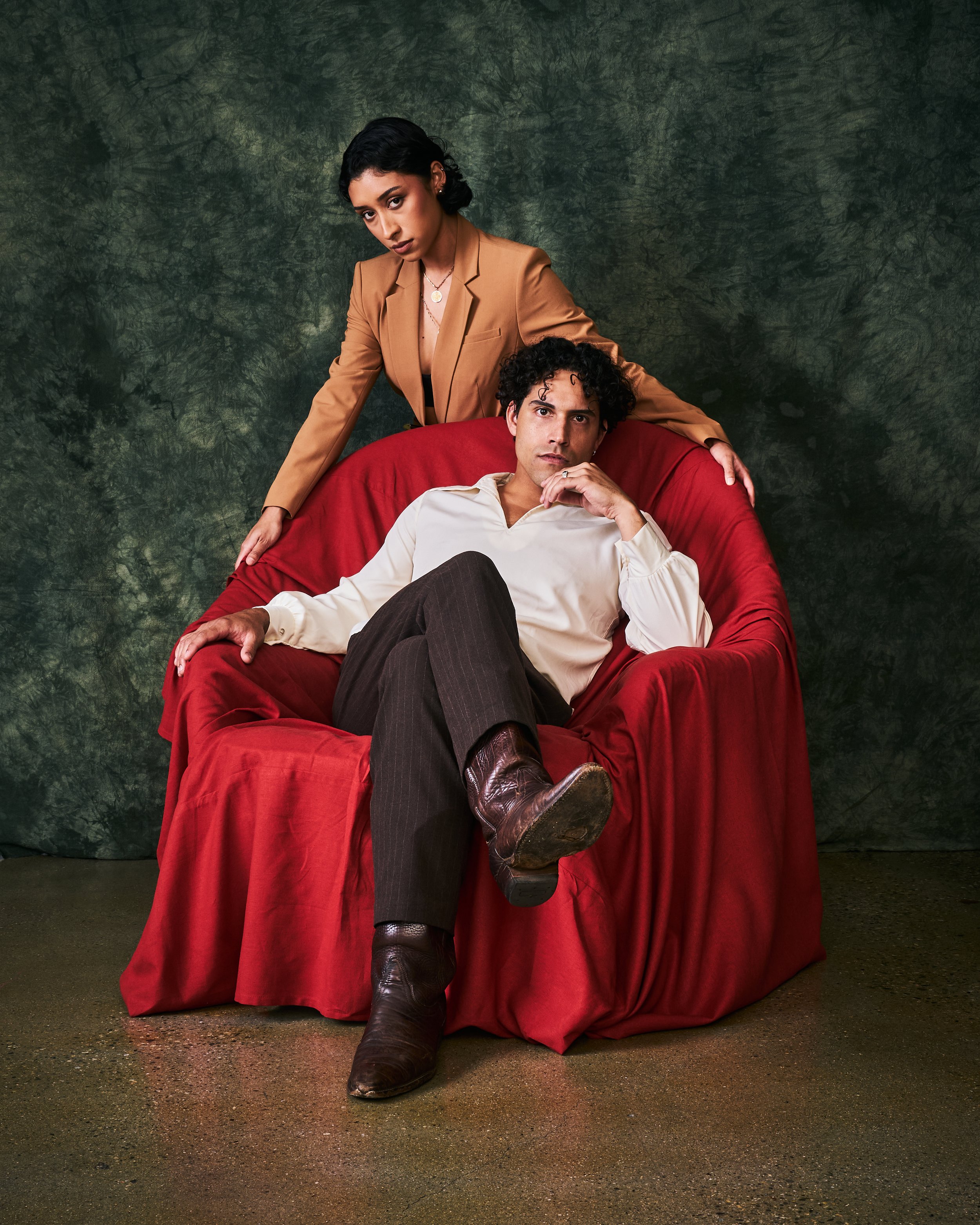 A woman in an orange blazer standing behind a man seated on a red velvet armchair, with a dark green textured background.