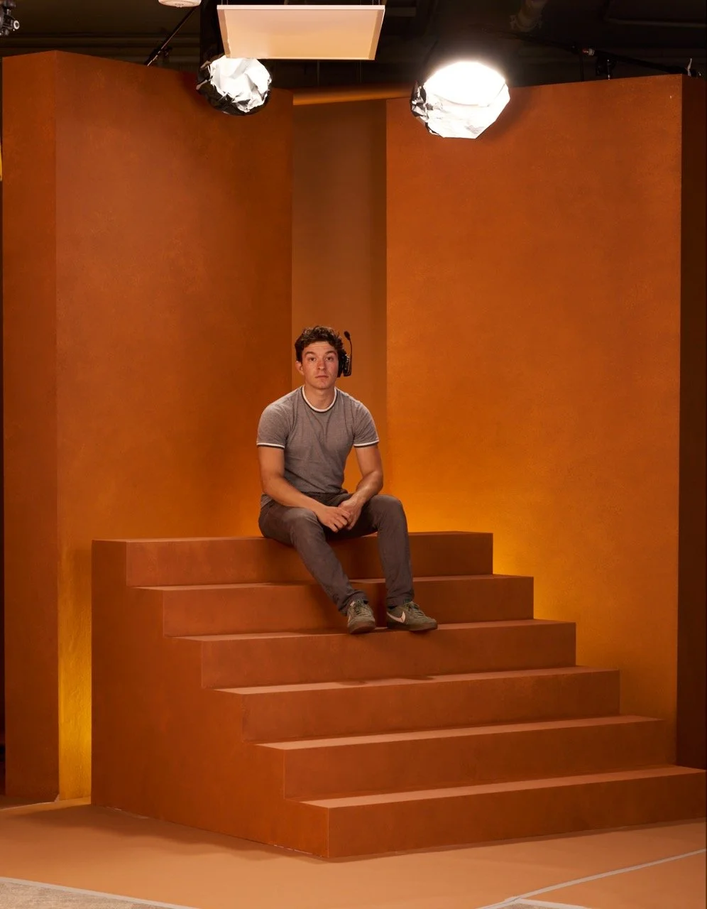 A young man sitting on a stepped stage in a studio with orange walls, wearing a grey T-shirt and headphones.