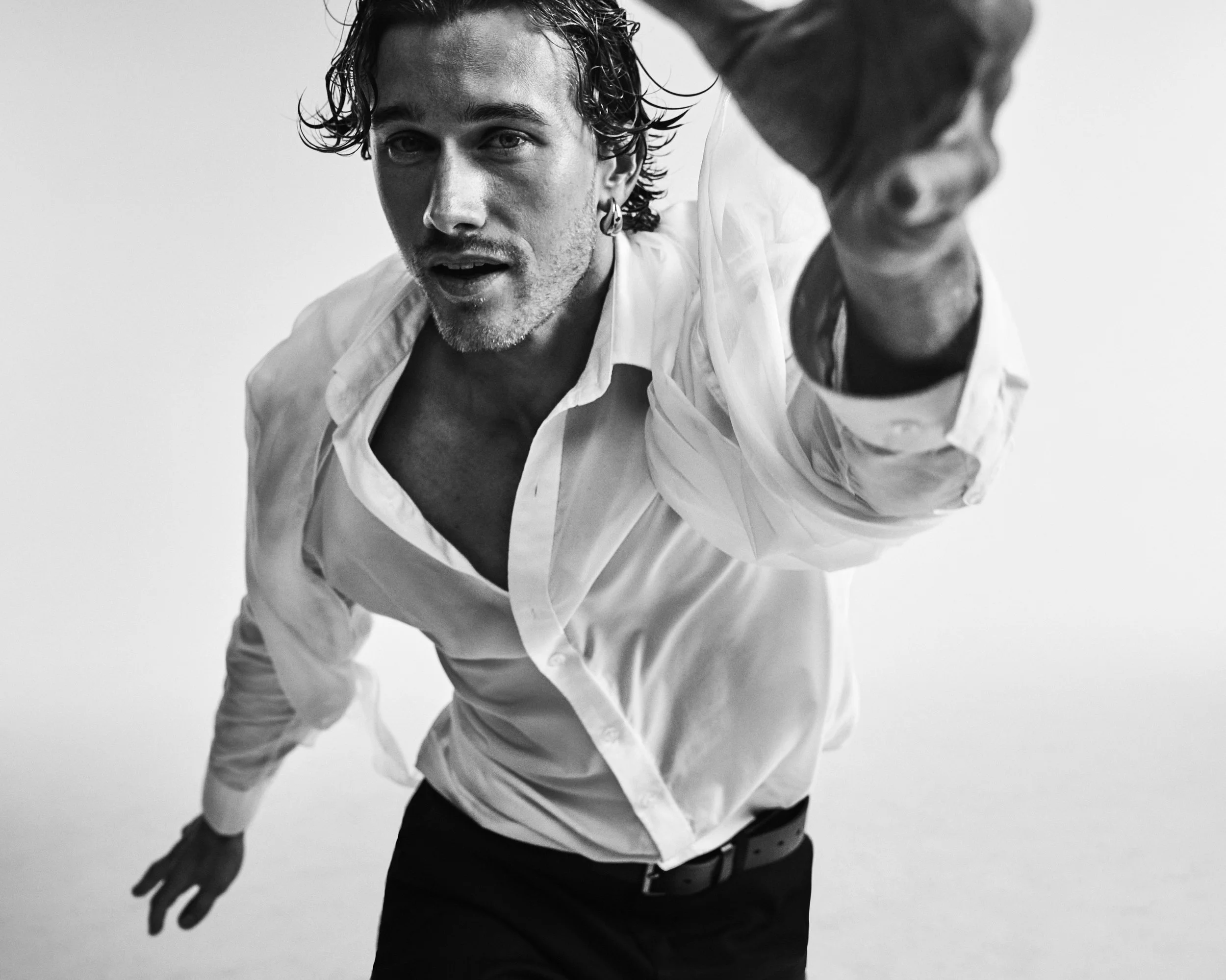 Black and white photo of a man with wet, curly hair, wearing a white shirt and black pants, reaching out towards the camera with his right hand.