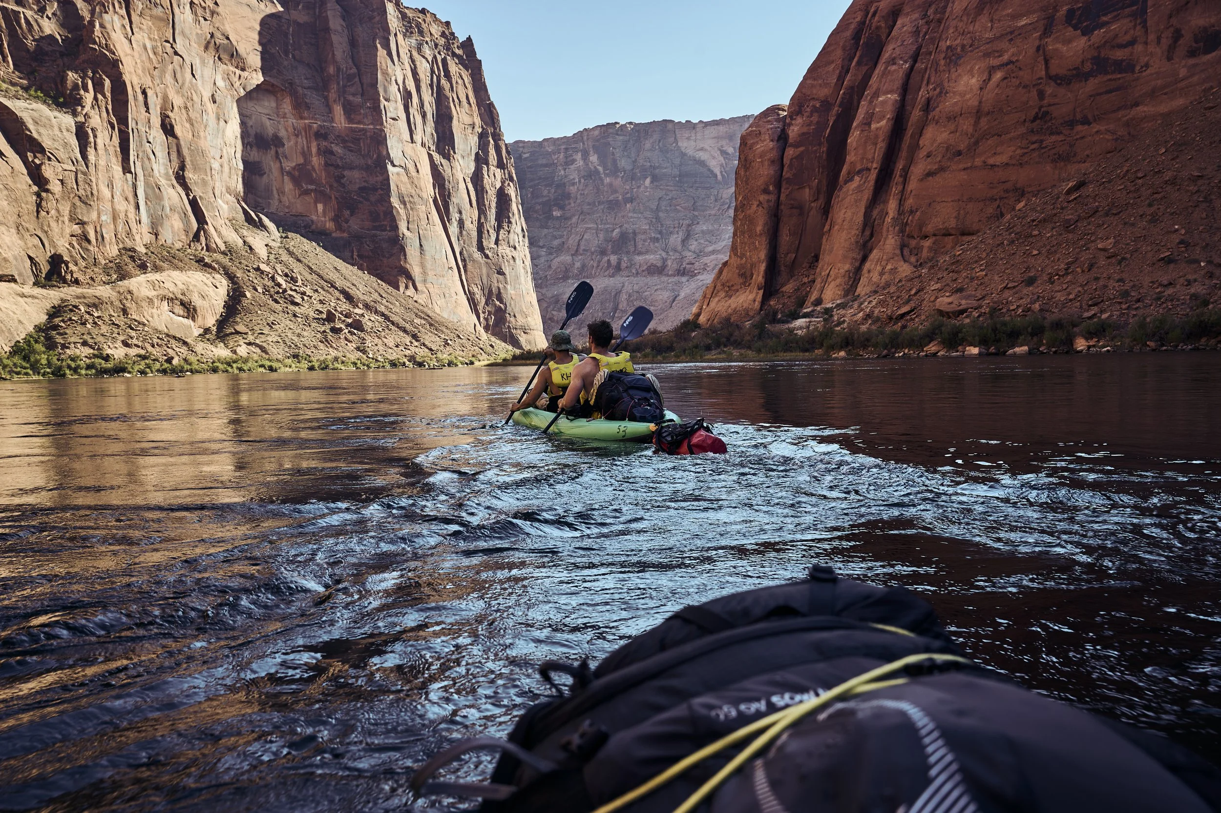 View from a kayak on a river flanked by towering red rock canyon walls with two people paddling.