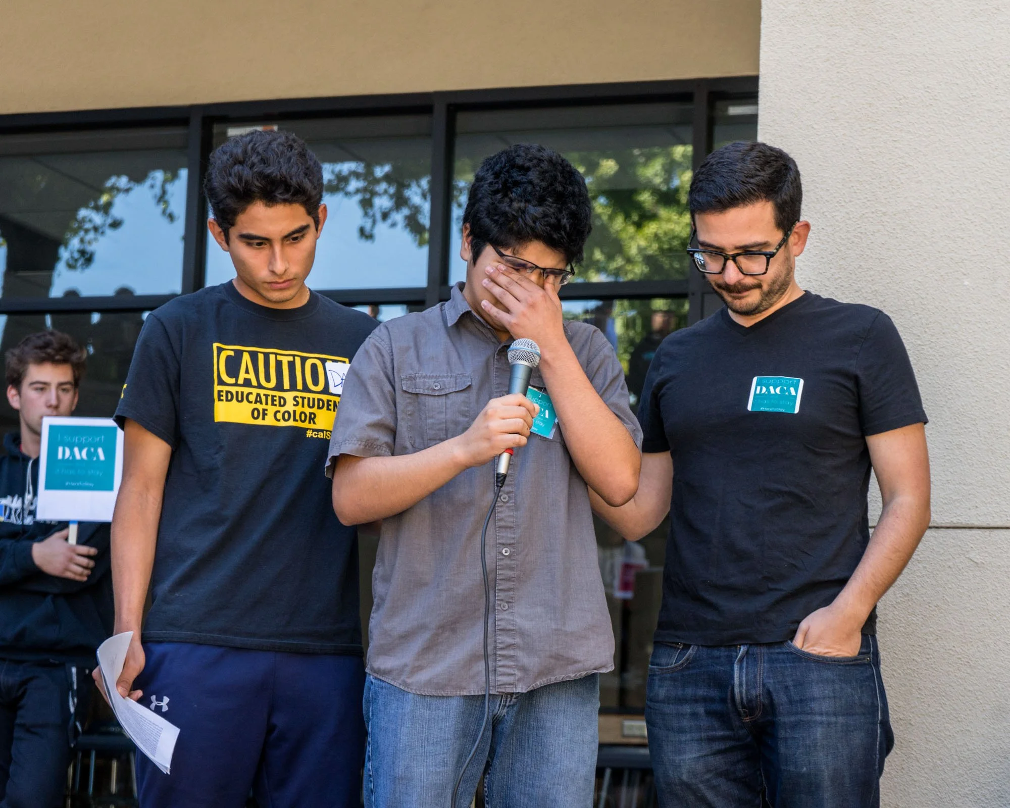 Three young men standing together outdoors, two of whom are wearing glasses. One man is holding a microphone and appears to be emotional, with his hand covering part of his face. The others are focused on him, standing close. Part of a group or event