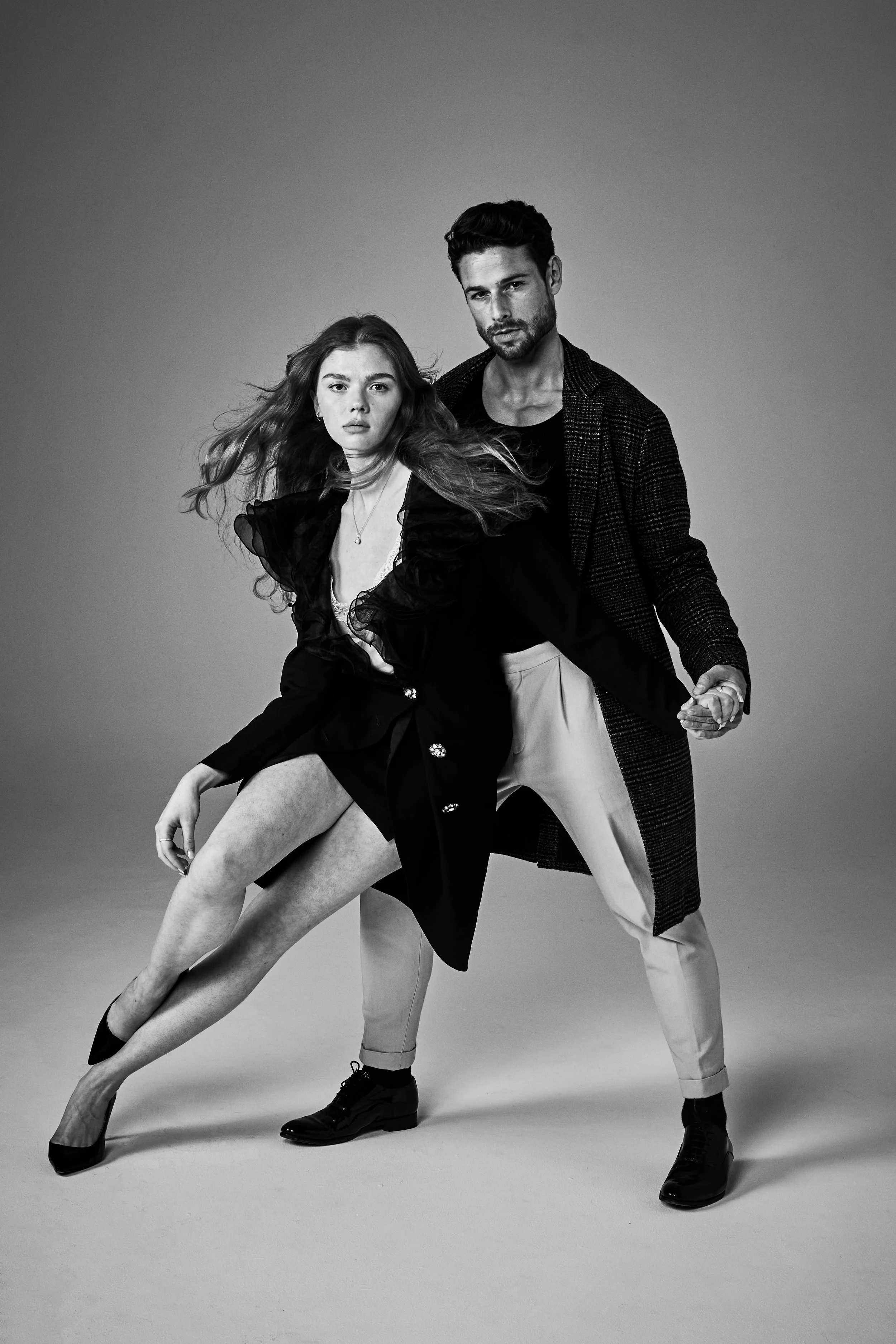 Black and white photo of a man and woman posing together against a plain background. The woman is sitting, leaning forward with her leg extended, wearing a dark dress with sheer sleeves and high heels. The man stands behind her, holding her hand, dre