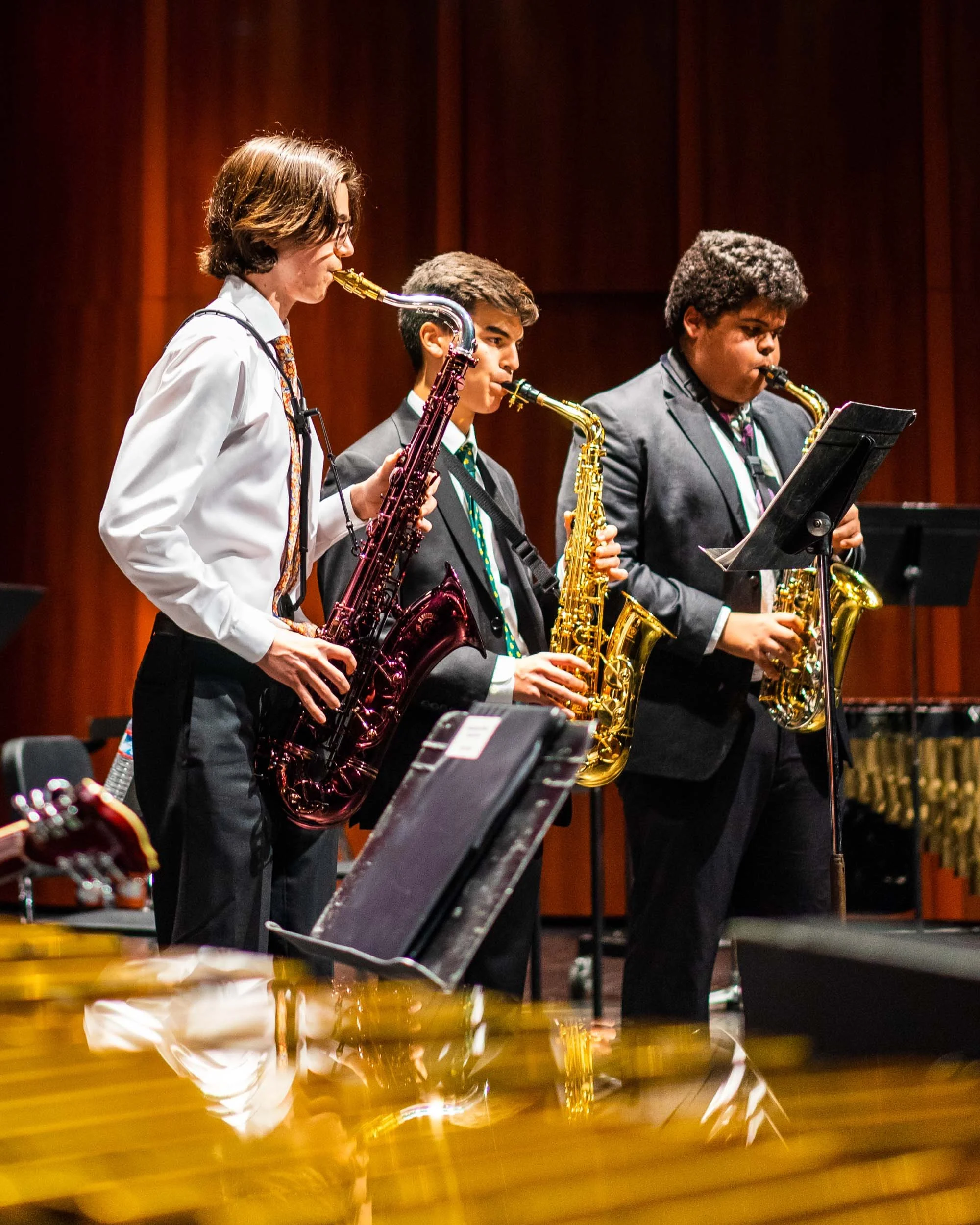 A group of four young musicians standing on stage playing saxophones, with a piano in the foreground and wooden wall panels behind them.
