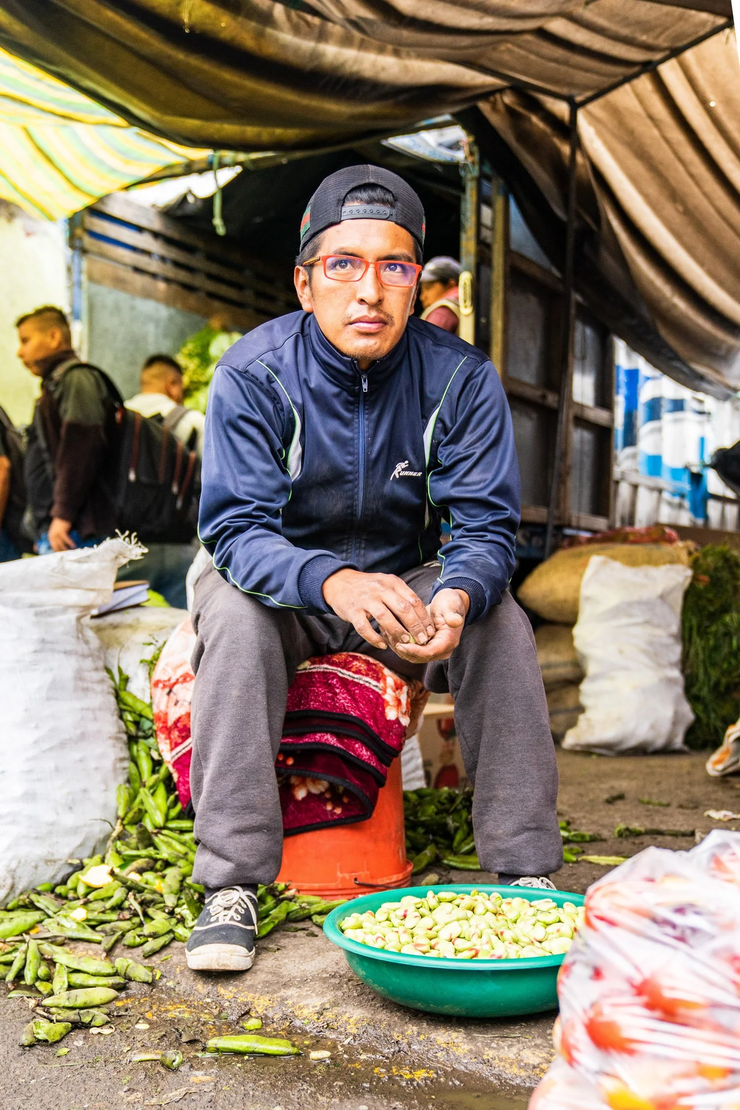 A man wearing glasses, a cap, and a navy jacket is sitting at a street market stall. Surrounding him are piles of green vegetables, including zucchini, and a large bowl filled with small, round light green vegetables. In the background, other market 