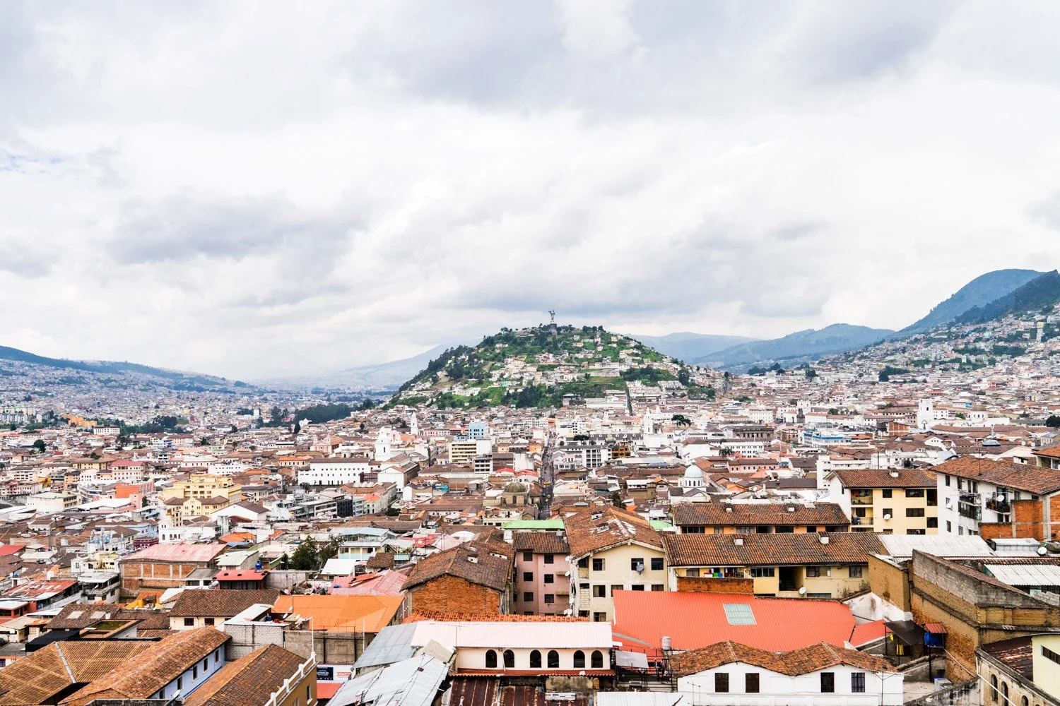 A panoramic view of a densely populated city with a green hill in the background and cloudy sky overhead.
