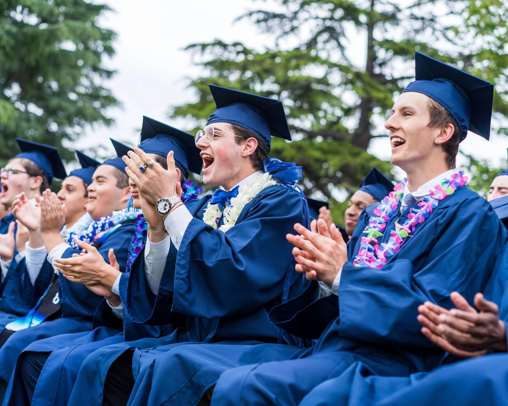 Group of young graduates wearing blue caps and gowns, clapping and celebrating outdoors during graduation ceremony.