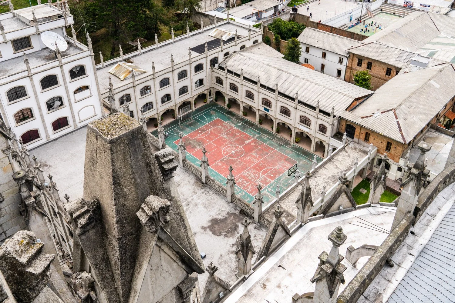 A view from a high vantage point looking down at a small outdoor basketball court surrounded by a historic church or cathedral with Gothic architectural elements, including stone spires and decorative pinnacles. The court is enclosed by a stone fence