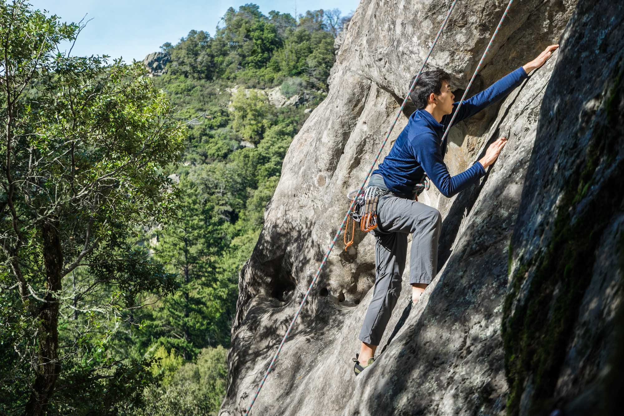 A person climbing a steep rock face outdoors, wearing climbing gear, with a forested hillside in the background.