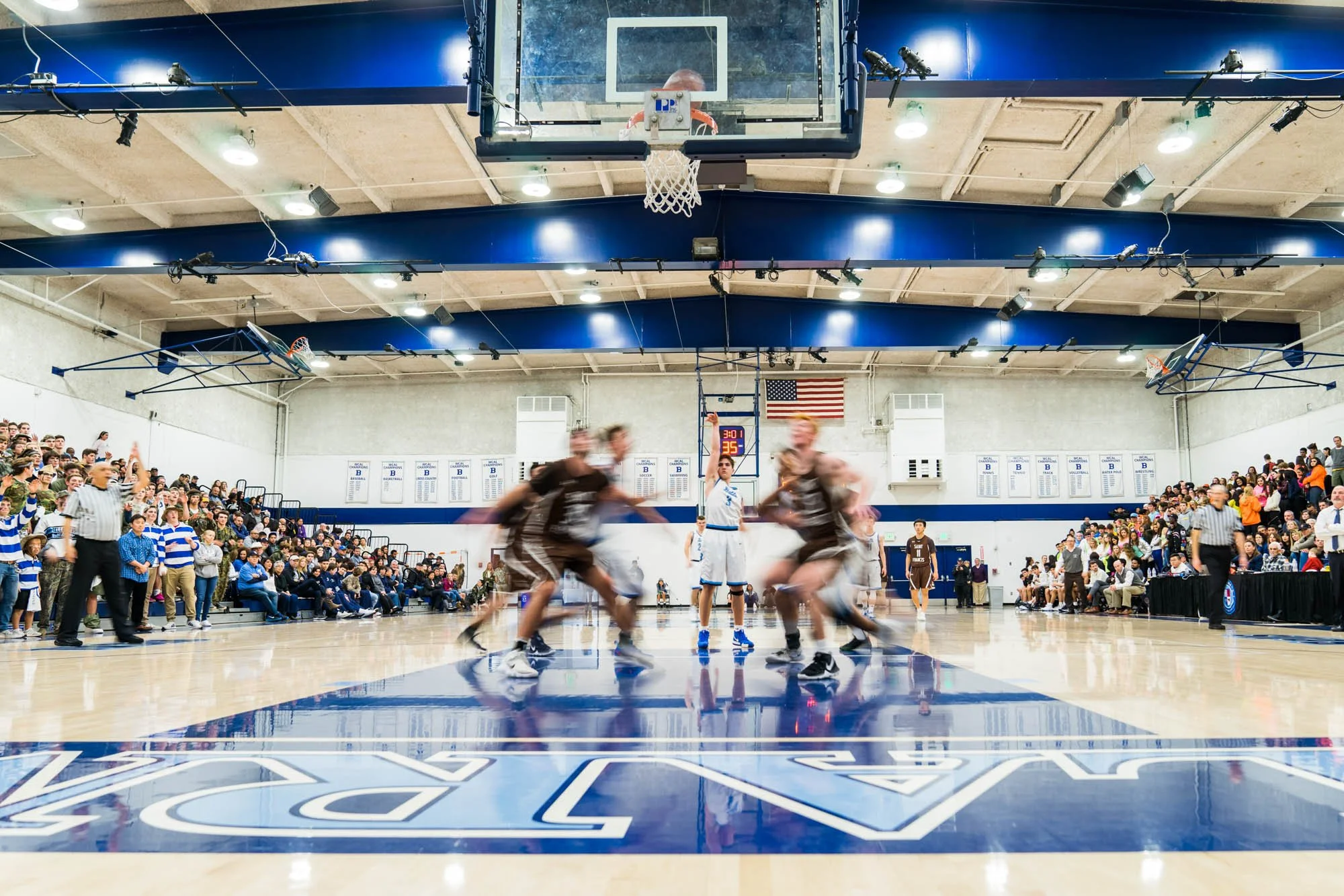 A basketball game in progress inside a gymnasium with spectators in the stands. Players are moving on the court, and the shot focuses on the backboard, hoop, and players below it. The gym has a crowd of students and spectators watching the game, with
