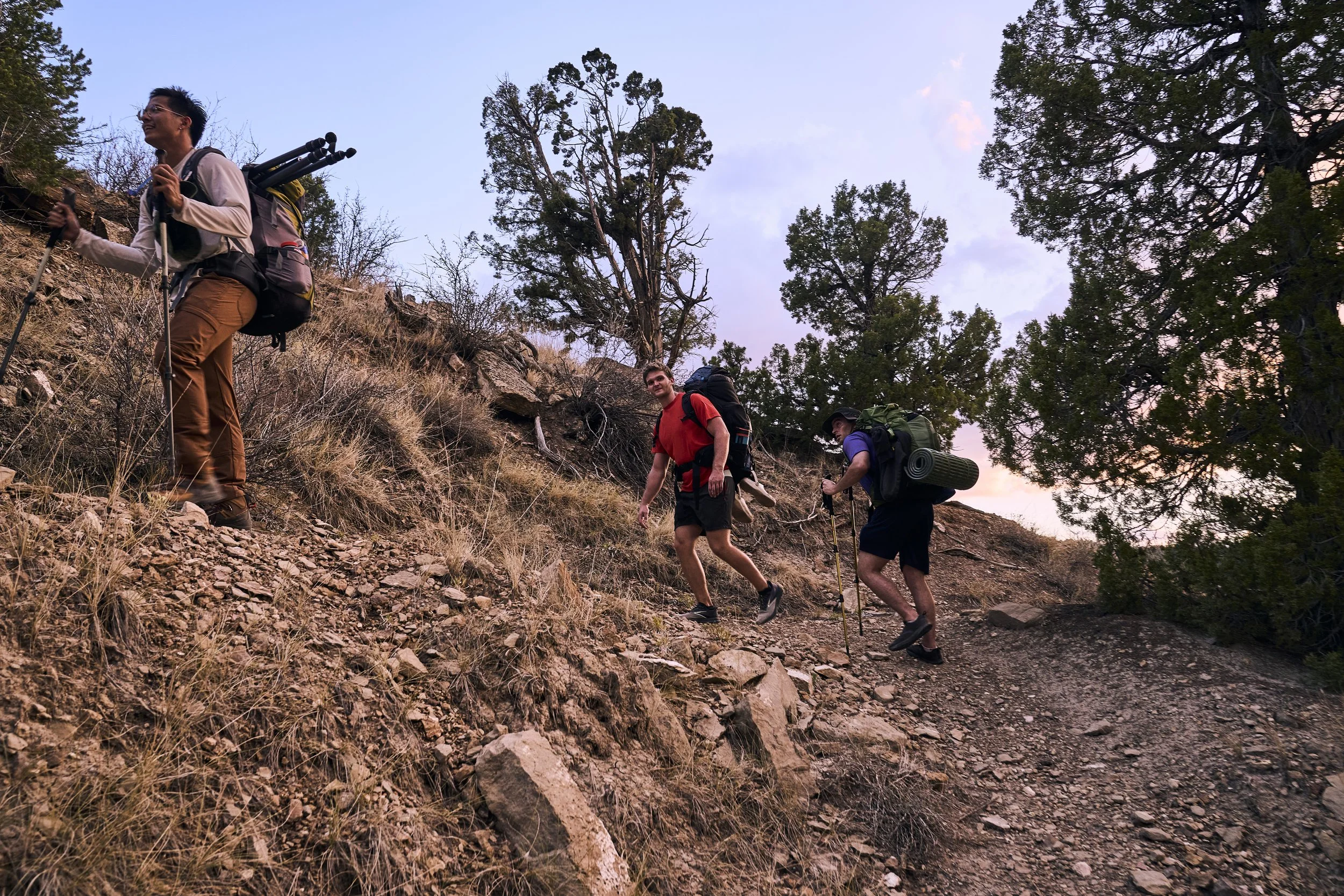 Three hikers with backpacks and trekking poles ascending a rocky trail through a forested area during sunset.