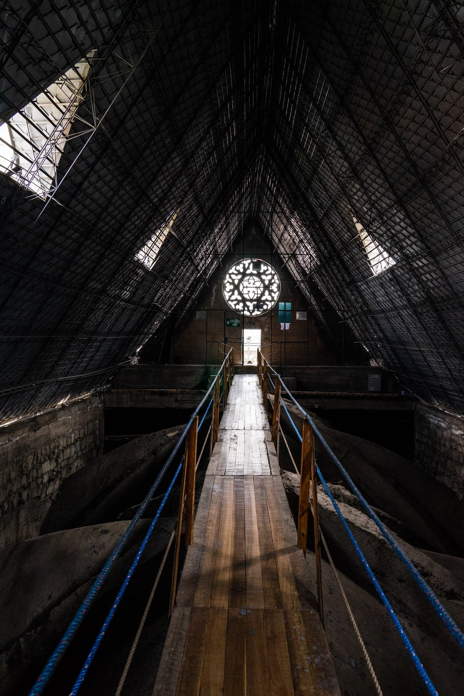 Inside a large, dark, metallic building with a high, triangular roof structure and a circular window at the end, featuring a wooden walkway with railings running through the center.