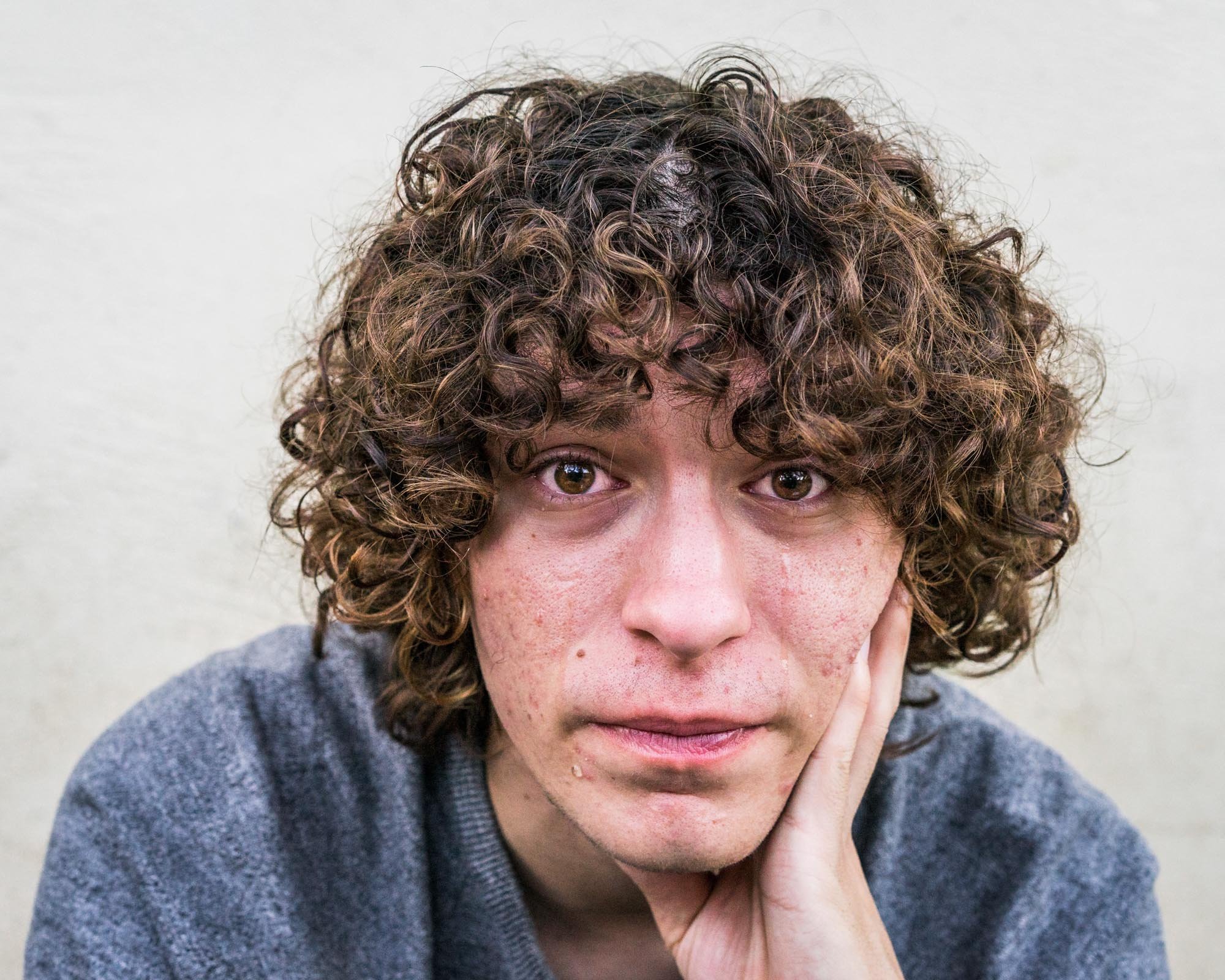 Close-up of a person with curly brown hair, resting their face on their hand, wearing a gray sweater.