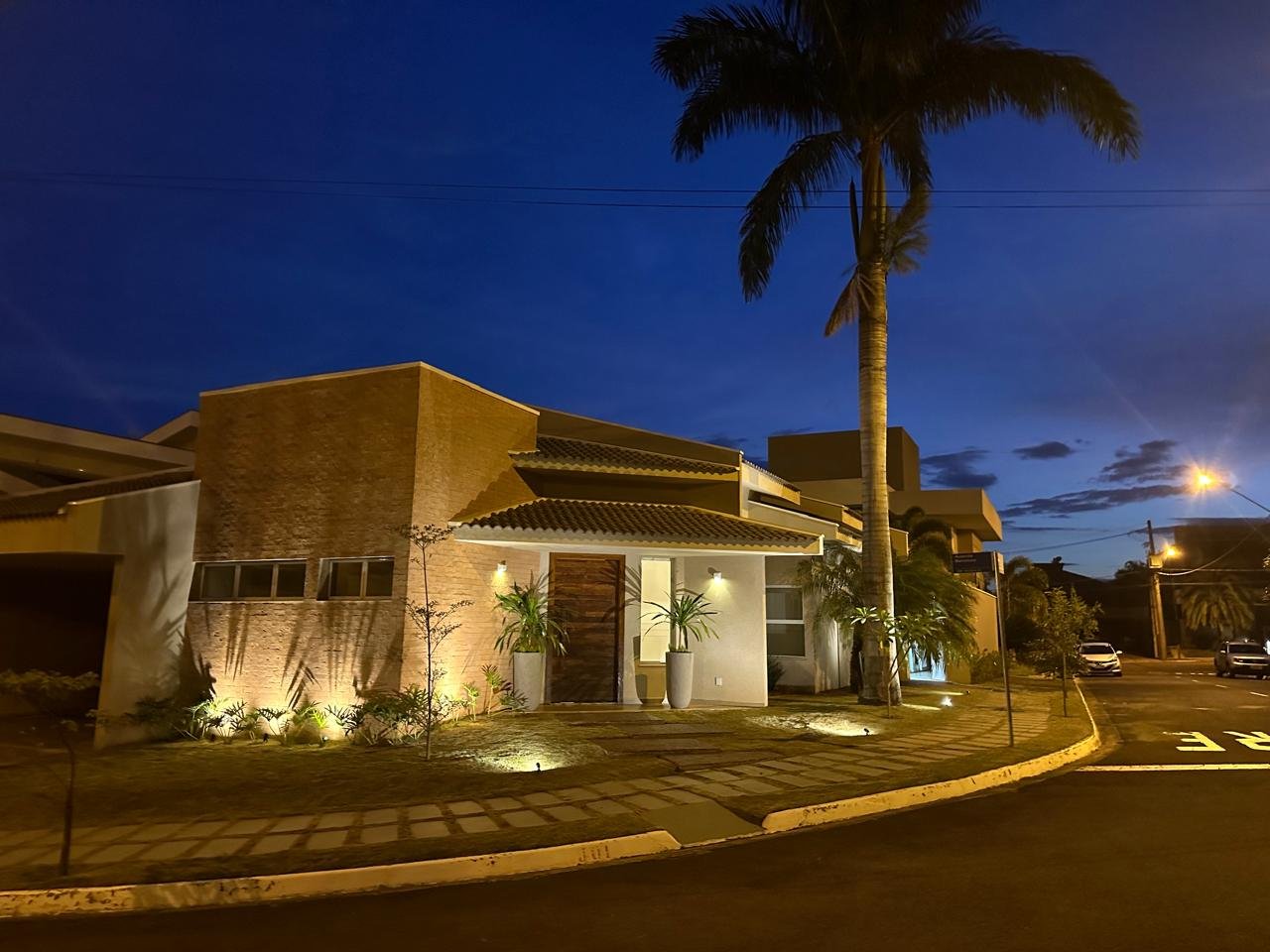 Casa moderna iluminada à noite, com árvore de palmeira na frente e céu escuro ao fundo.