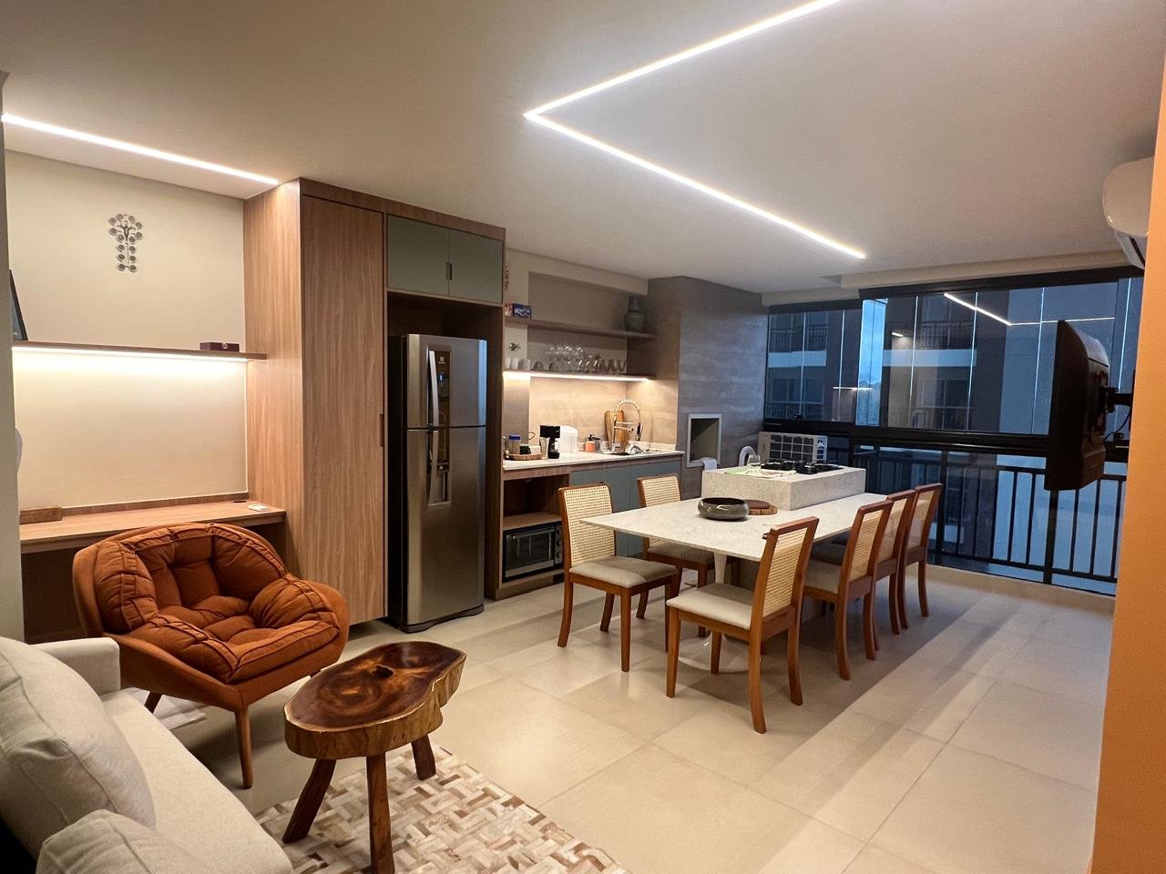 Imagem de uma sala de jantar e cozinha em um apartamento moderno, com móveis de madeira, uma mesa de jantar com seis cadeiras, cozinha com geladeira de inox e eletrodomésticos, janelas amplas com vista para outros prédios, iluminação de LED no teto, ambiente bem iluminado e decorado de forma contemporânea.