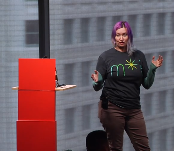 A woman with purple and gray hair giving a presentation in a conference room with large windows. She wears a black shirt with green text and a yellow starburst, brown pants, and has a microphone attached to her waist.