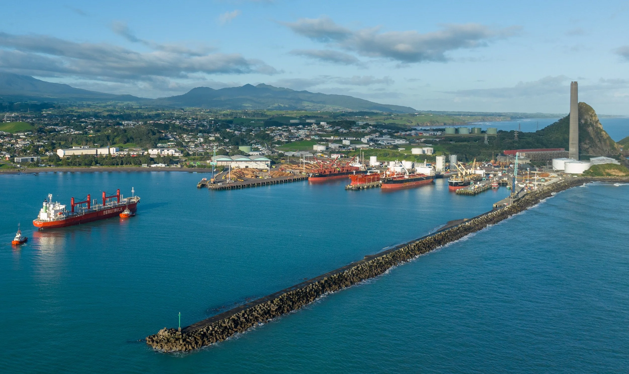 An aerial view of a harbor with several ships docked, a long breakwater extending into the water, industrial buildings, and a cityscape with mountains in the background under a partly cloudy sky.