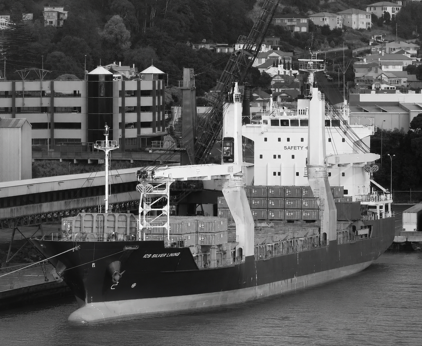 A large cargo ship docked at a port with containers on board, a crane overhanging the ship, and buildings on a hillside in the background.