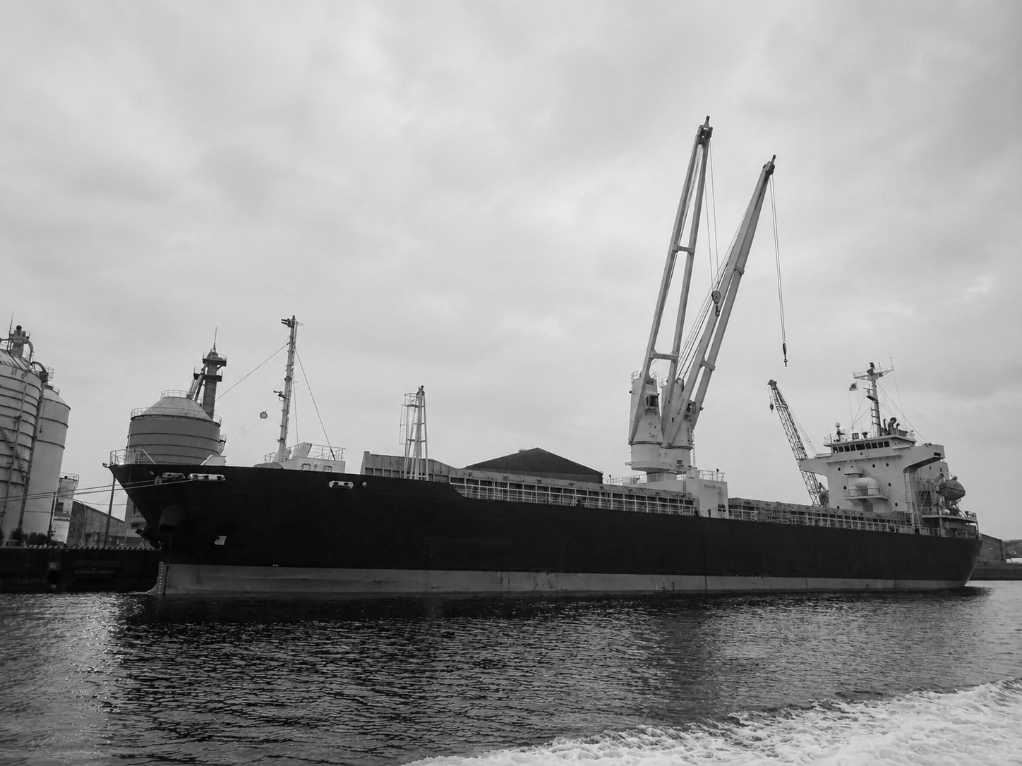 Black cargo ship docked at port with cranes and industrial buildings in the background, black and white photo.