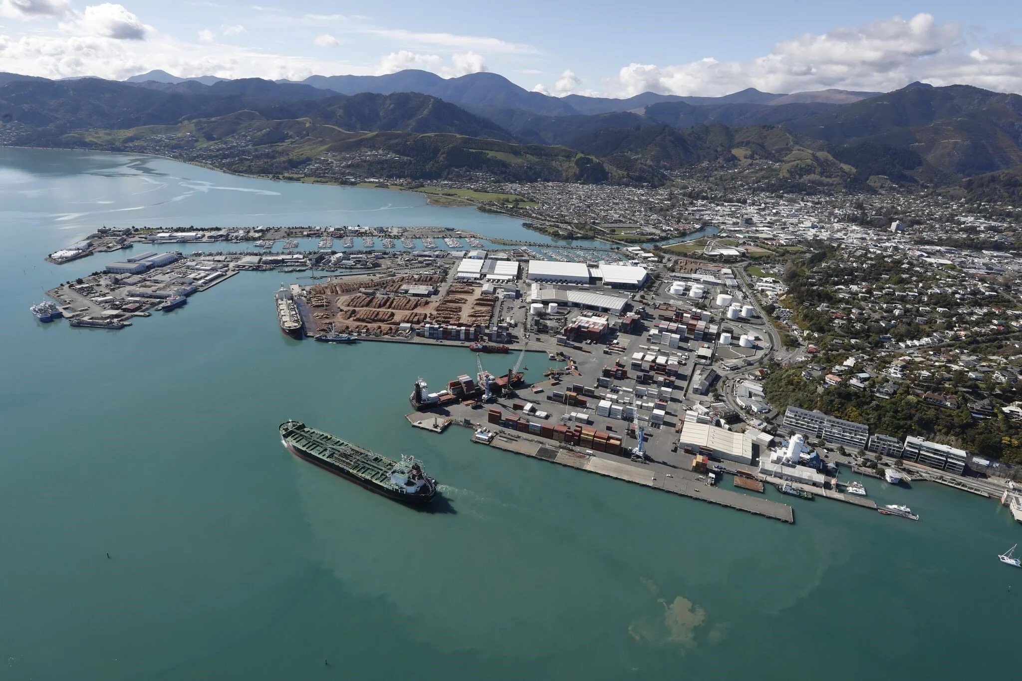 An aerial view of a harbor with ships, containers, industrial buildings, and warehouses. There are mountains in the background and a cityscape along the shoreline.