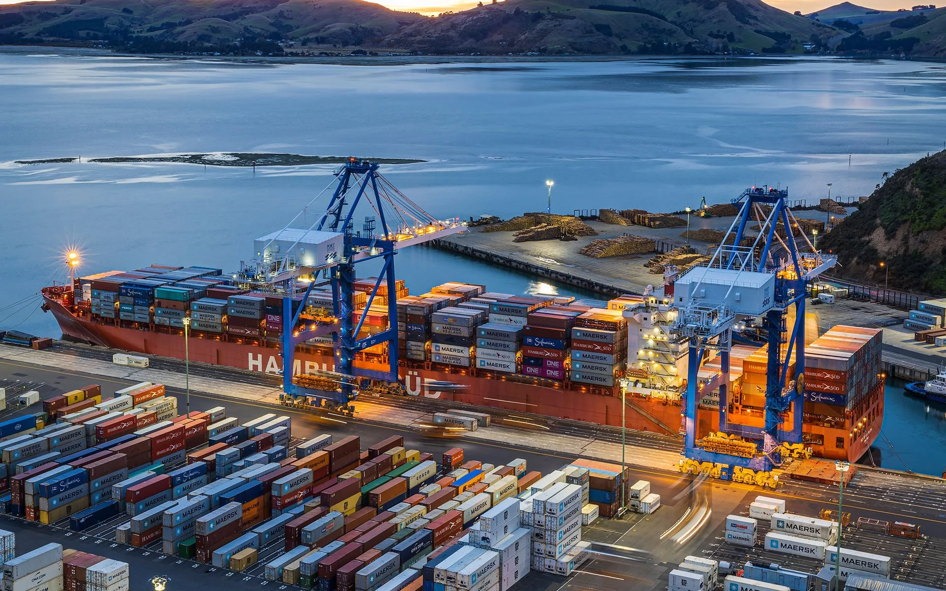 A large cargo ship docked at a port with numerous shipping containers, cranes, and water in the background, during twilight.