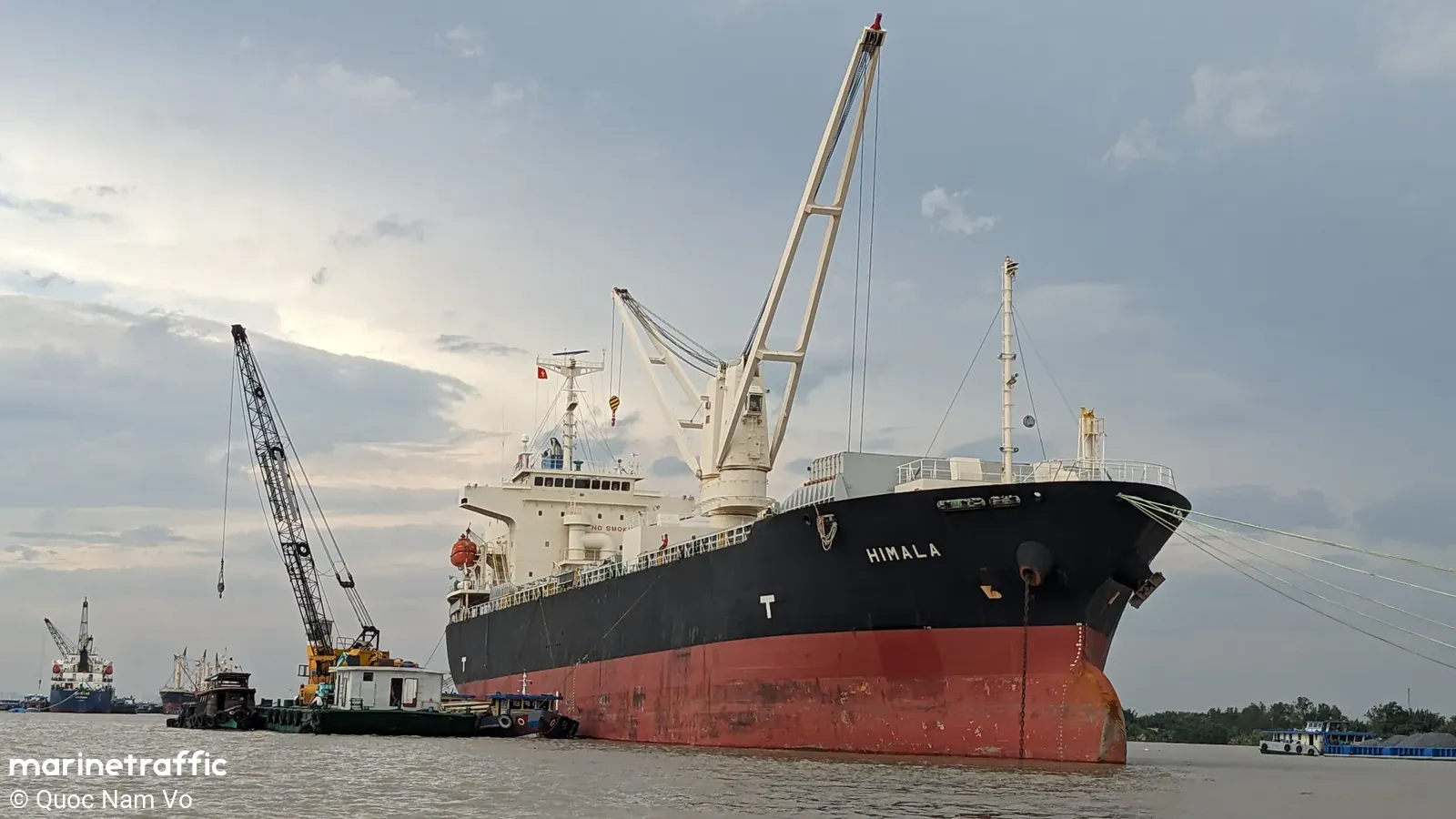 Large black and red tanker ship named Himalia docked at a port with cranes and tugboats nearby under cloudy sky.
