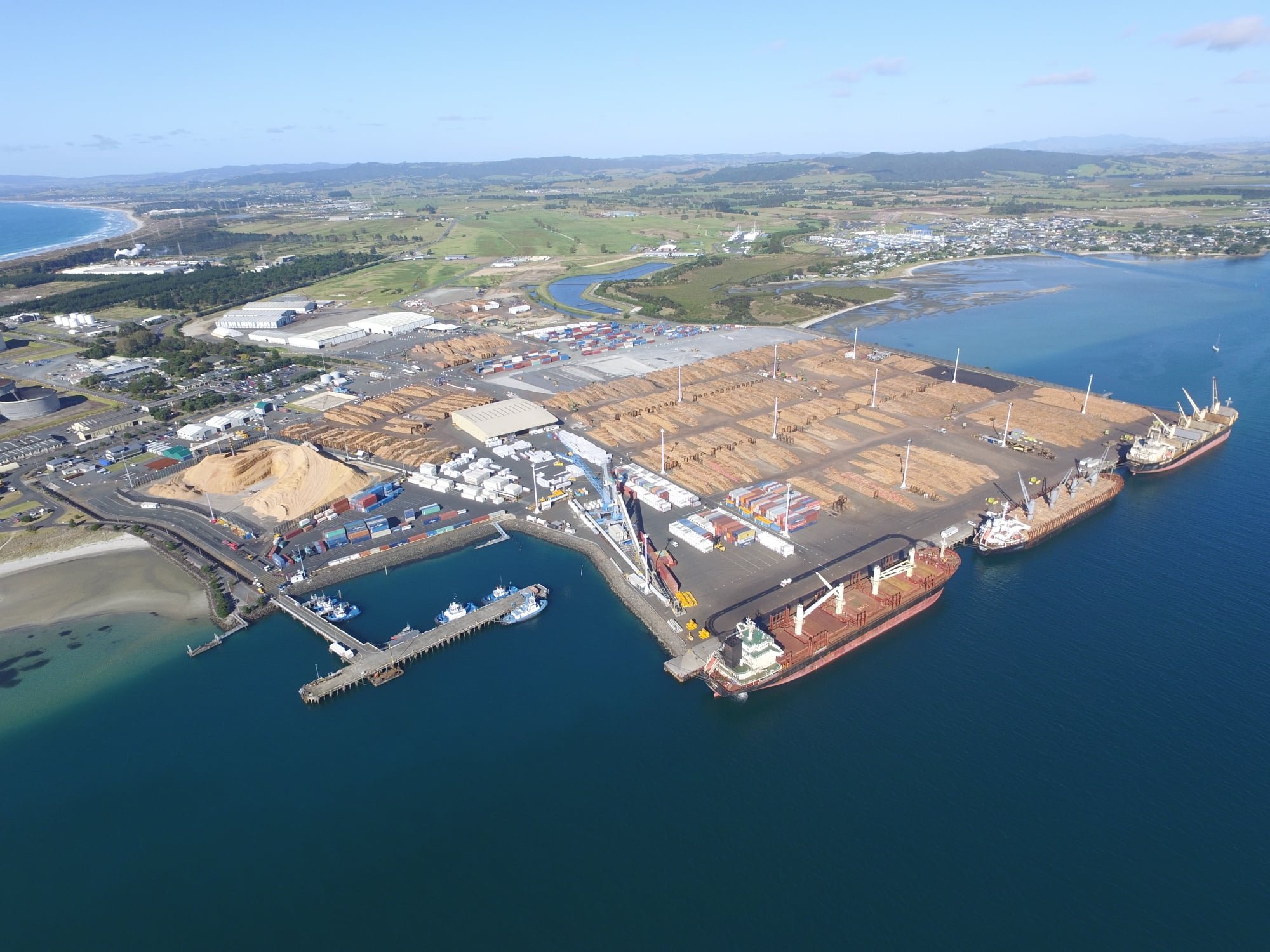 Aerial view of a harbor with cargo ships, containers, and warehouses, adjacent to a body of water with a coastal town and green landscape in the background.