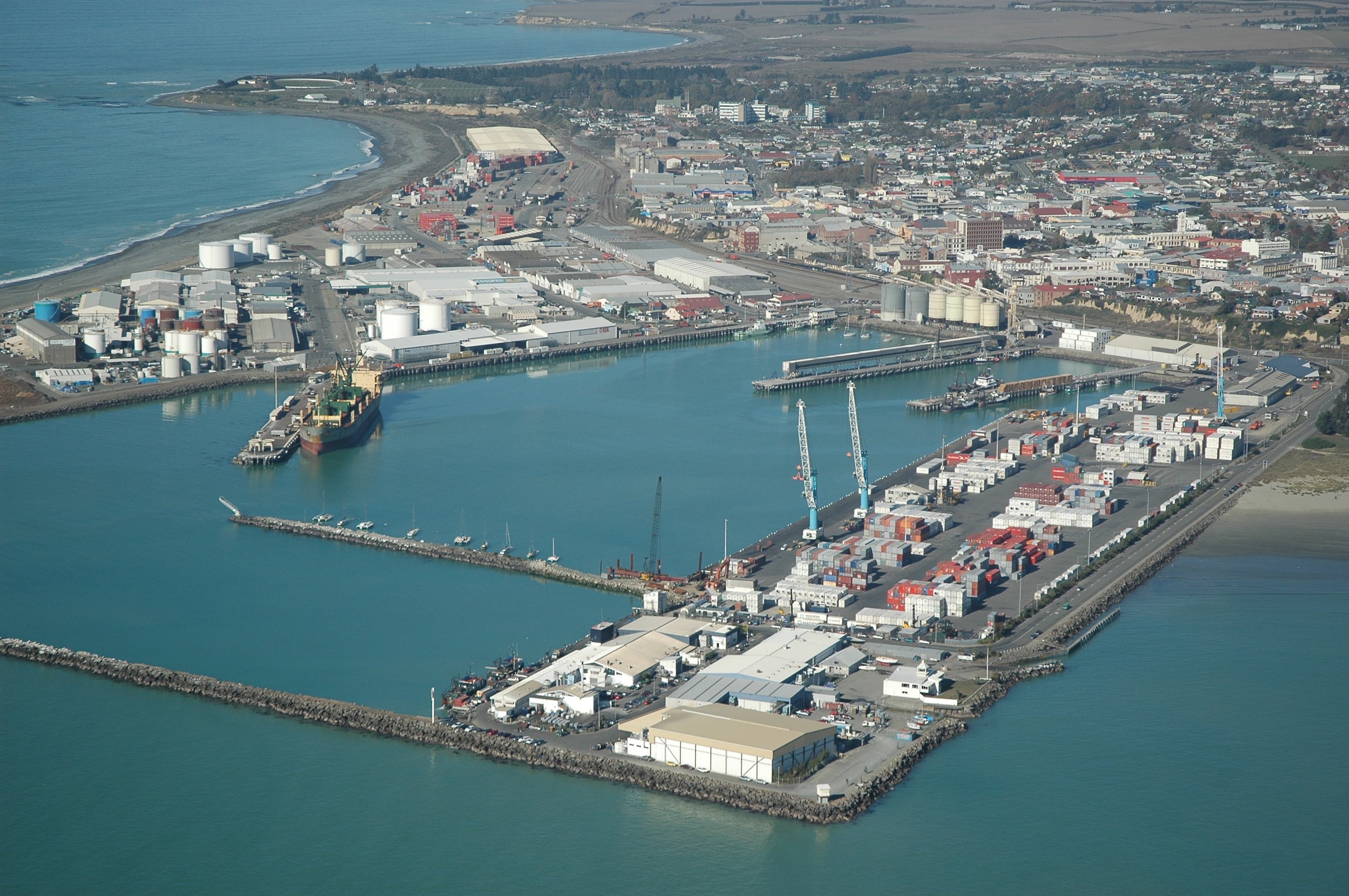 Aerial view of a harbor with cranes, shipping containers, warehouses, and a ship docked at the pier. Beyond the harbor, there is a city with numerous buildings and industrial structures near the coastline, with a beach and ocean in the background.