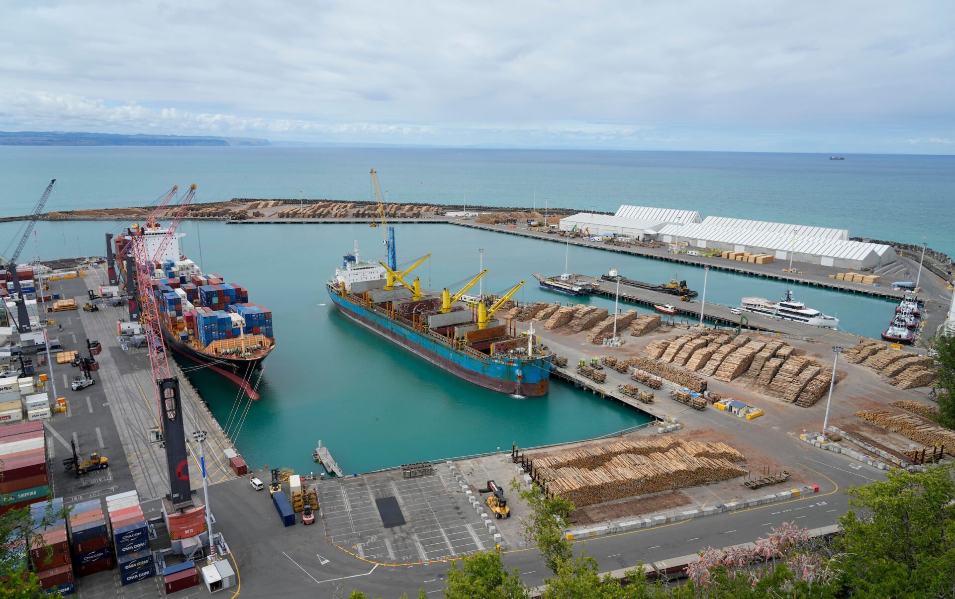 A busy port with ships, cargo containers, cranes, and warehouses on the ocean and cloudy sky in the background.