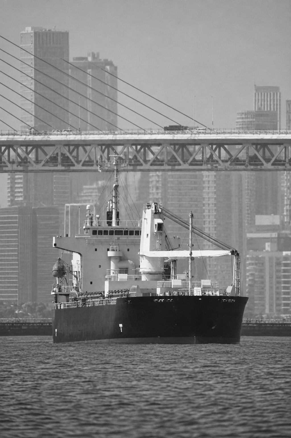 Black and white photo of a large cargo ship sailing on water with a city skyline and bridge in the background.