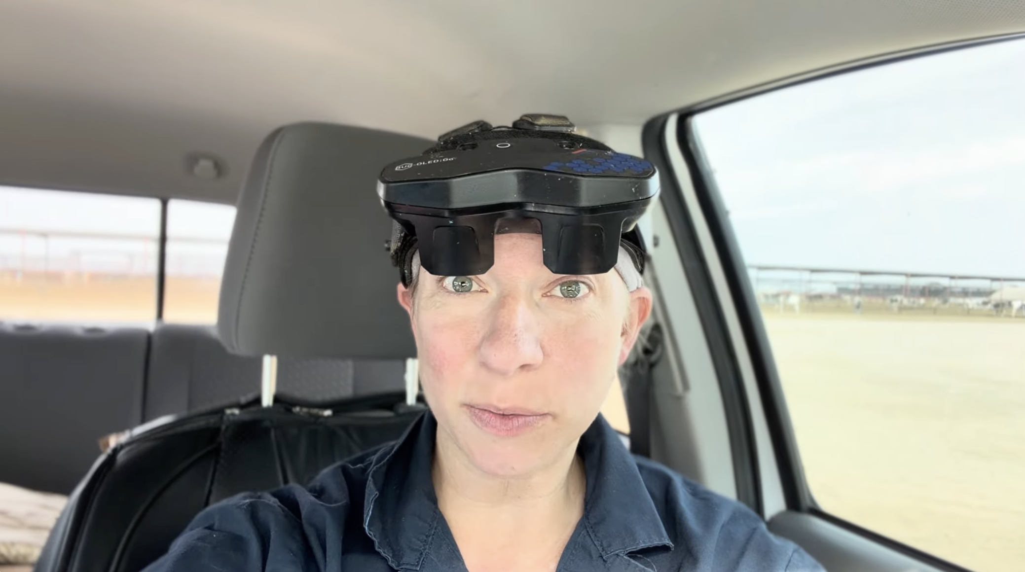 A woman with short light-colored hair and green eyes sitting in the back seat of a vehicle, wearing a head-mounted device, looking at the camera, with a field visible through the window behind her.