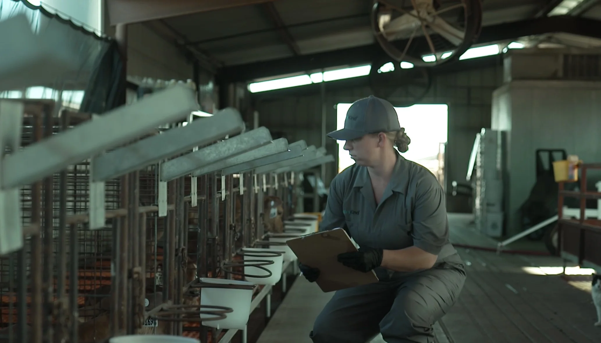 A person wearing a gray uniform and cap sits in a warehouse or industrial setting, holding a clipboard and taking notes, with metal racks and equipment around.