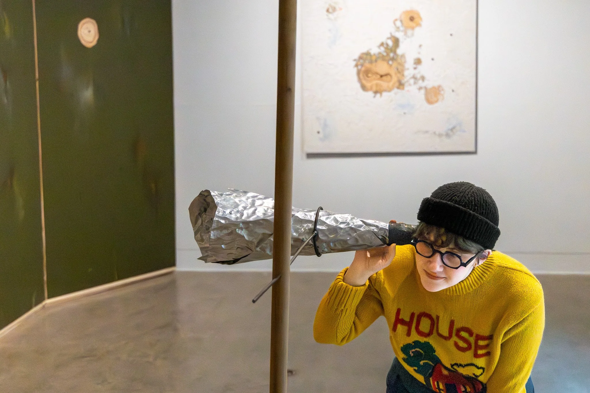 A young boy with glasses and a black knit cap is crouching and looking intently at a science experiment setup that includes a cardboard tube, aluminum foil, and wires, with a colorful wall art in the background.