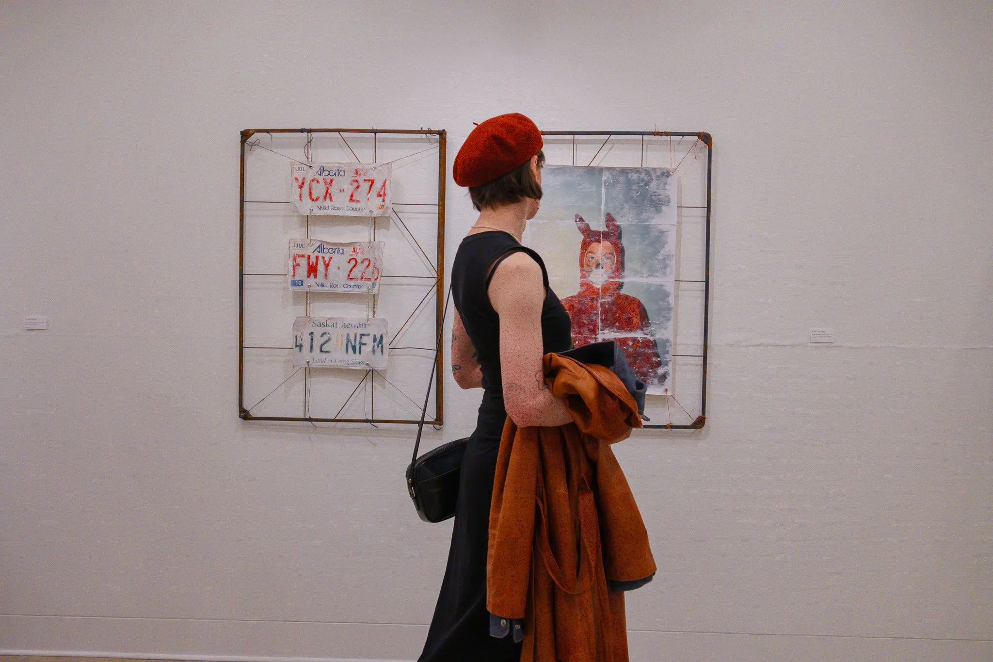 A woman with short brown hair wearing a red beret and a black sleeveless top stands in front of artwork in an art gallery. She holds a brown coat and a backpack, and the artwork behind her features a portrait of a person with animal ears and a face m