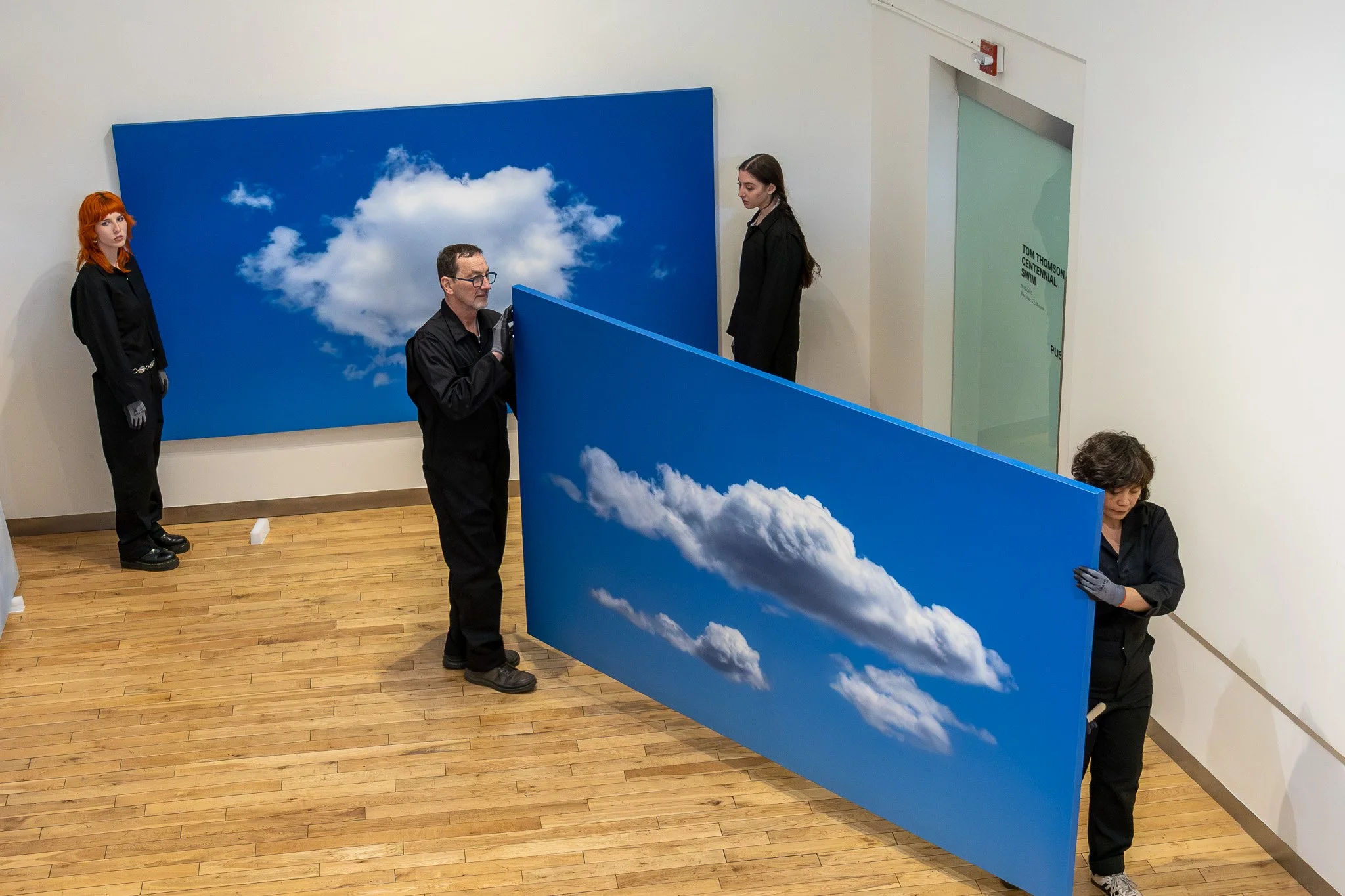 Four people setting up a large blue panel with a cloud sky print in an art gallery. One person with orange hair stands to the side, another man with glasses is holding the panel, and two women are preparing to position it, all wearing black clothing.