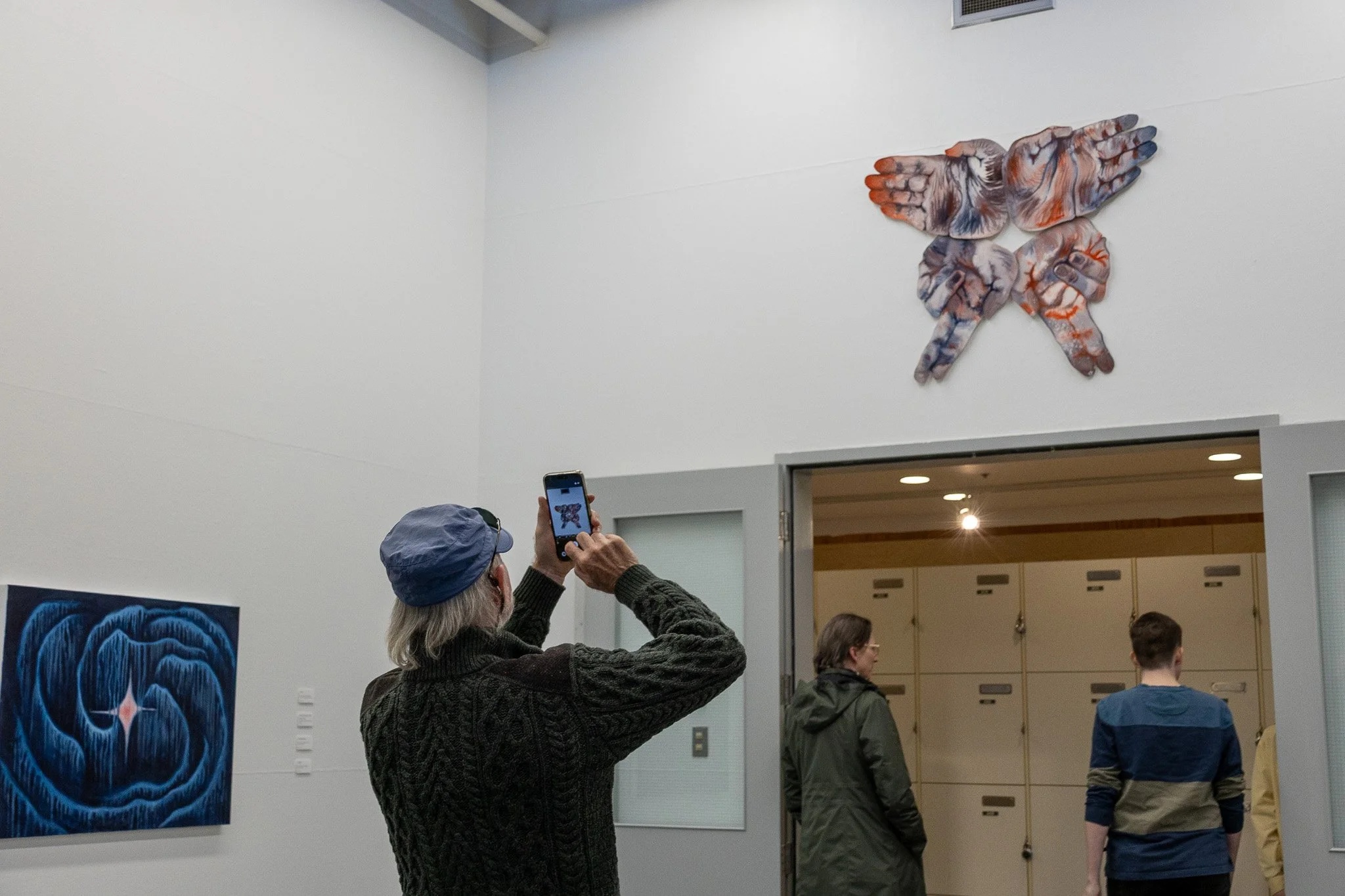 A woman taking a photo of wall art that features a 3D sculpture of two hands touching, in an art gallery with people walking and artwork on the walls.