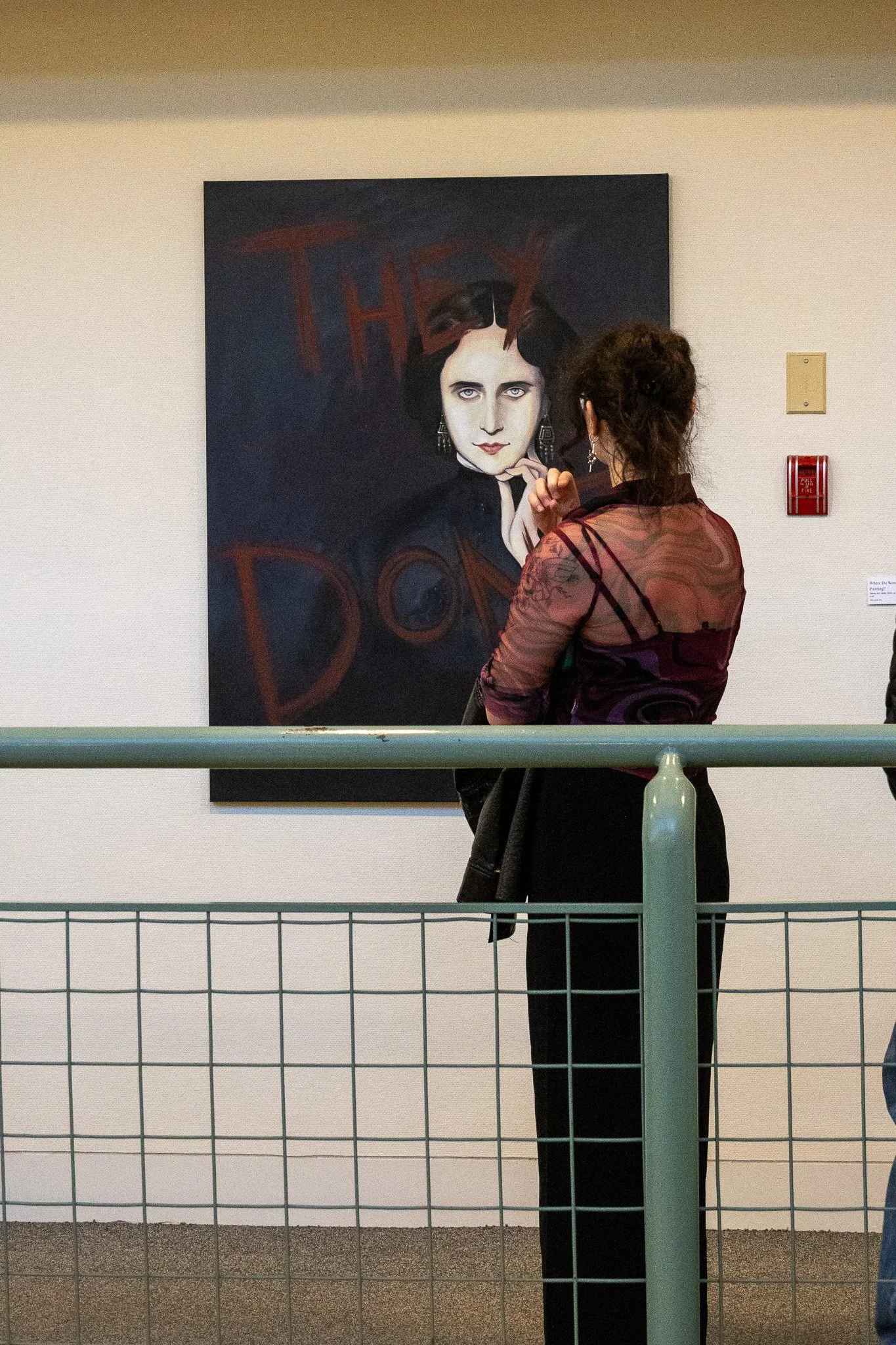 A woman with dark hair styled in a messy bun stands with her back to the camera, observing a portrait of a woman with pale skin, dark hair, and dark eyes, dressed in black and wearing earrings, in an art gallery. The gallery has a beige wall with a f