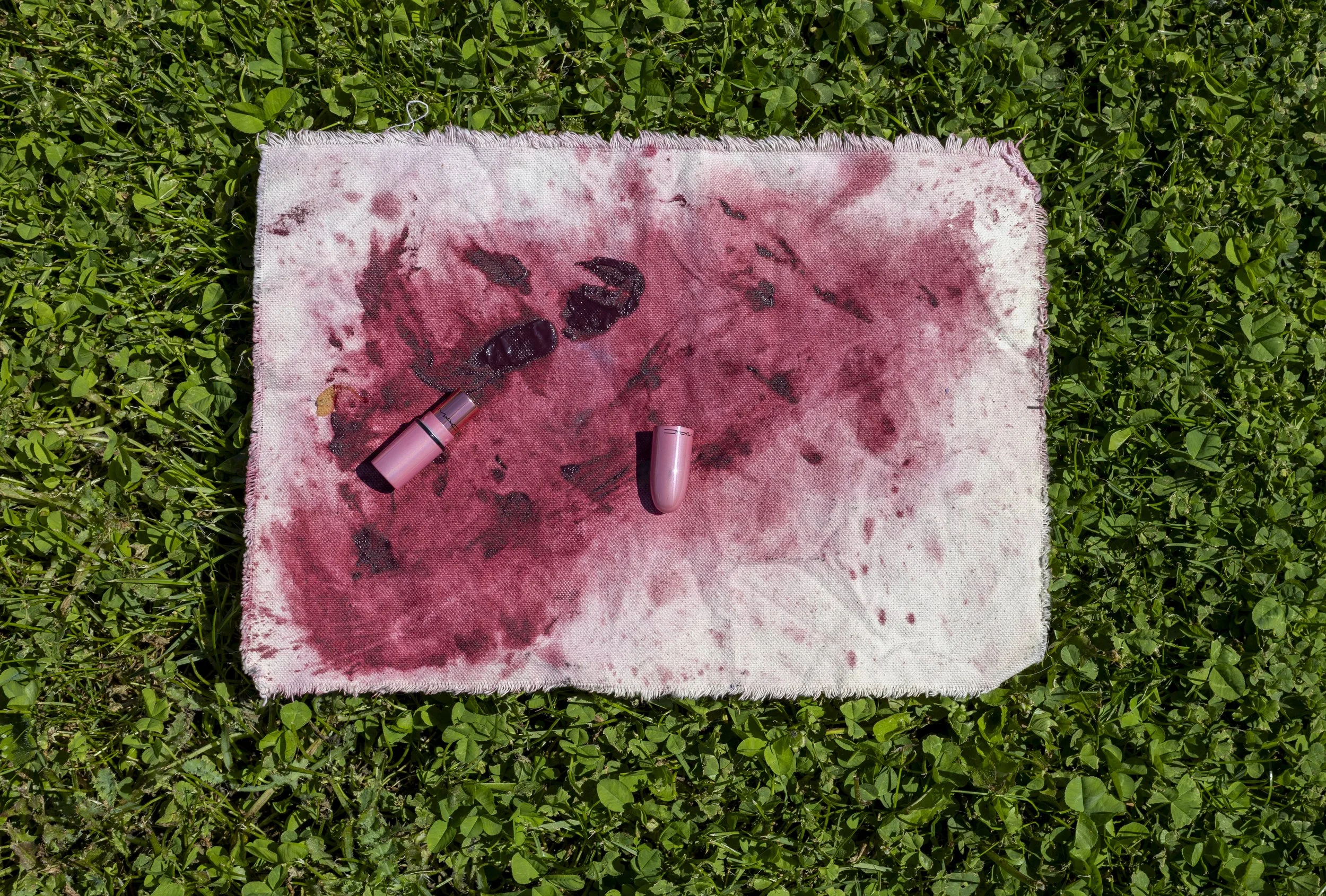 A bloodstained cloth with a large bloodstain and several smaller bloodstains on grass, with two pink lipstick tubes placed on the cloth.